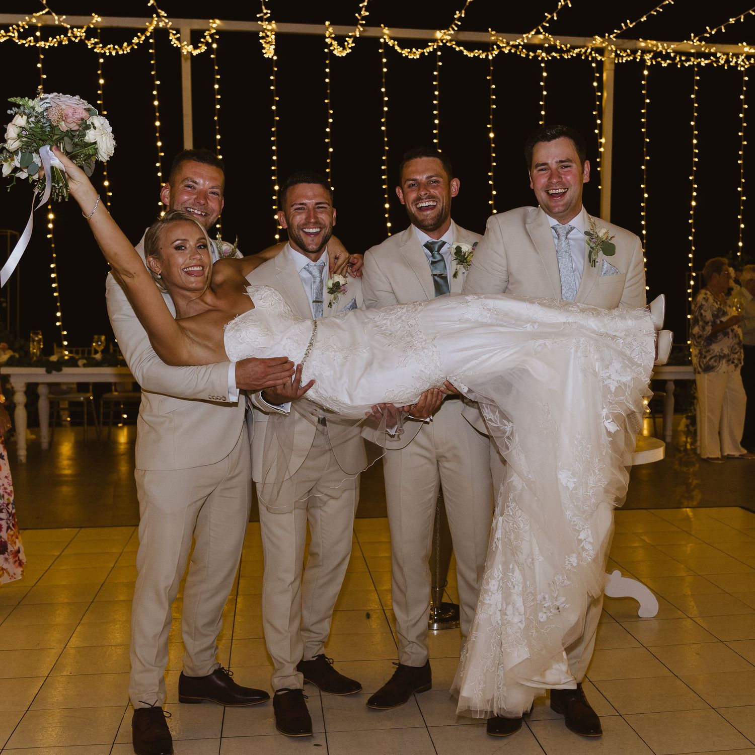 Wedding party lifts a bride in a white dress while groomsmen wear light beige suits under twinkling string lights.