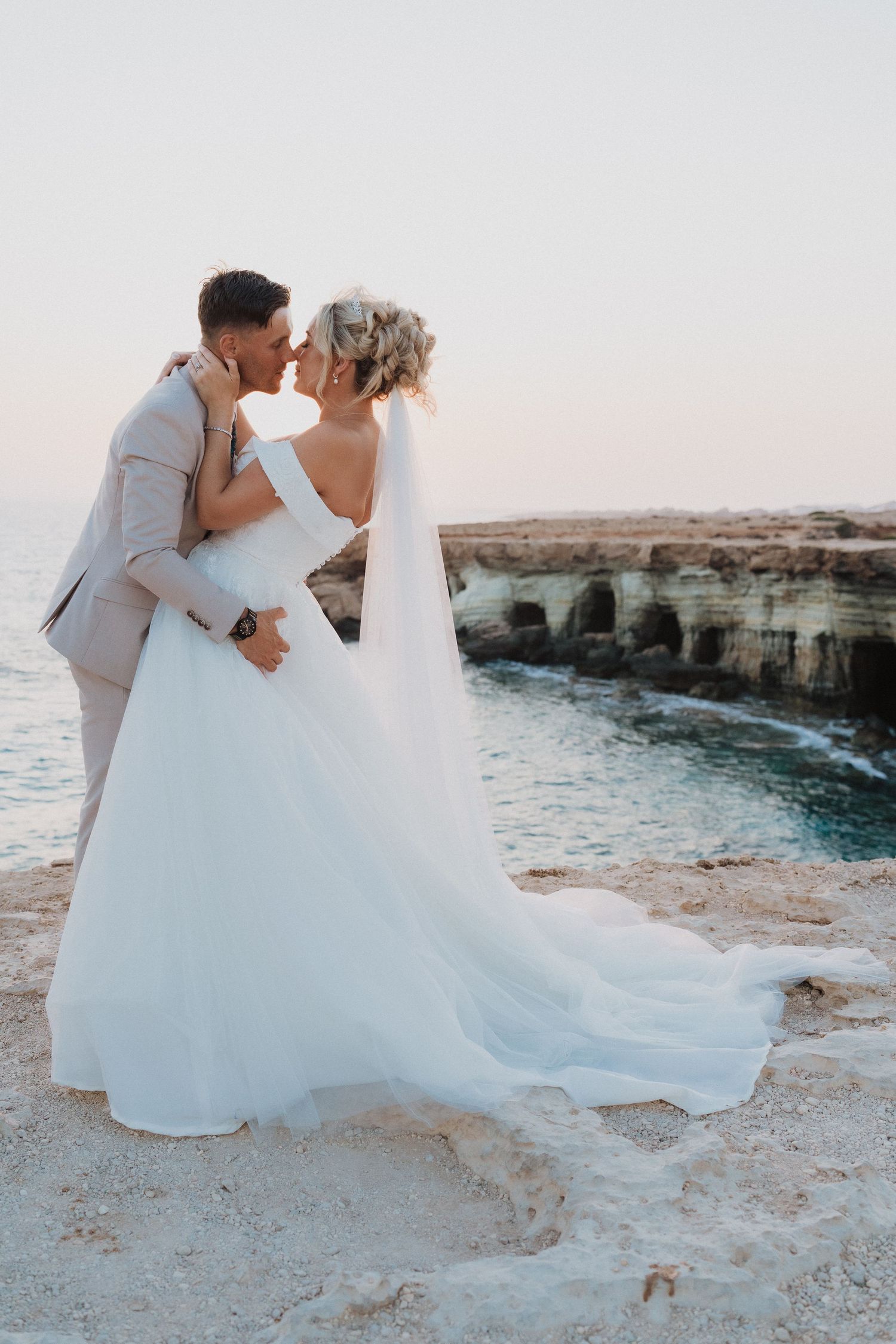 Couple sharing an intimate moment on coastal cliffs during sunset in formal wedding attire.