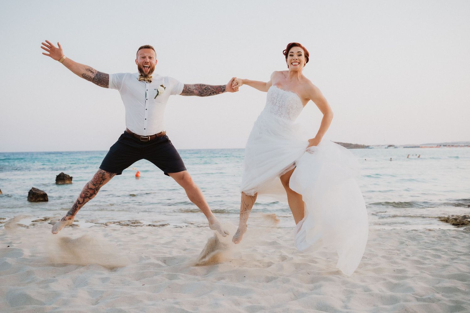Playful beach wedding photoshoot with couple jumping on sandy shore near ocean.