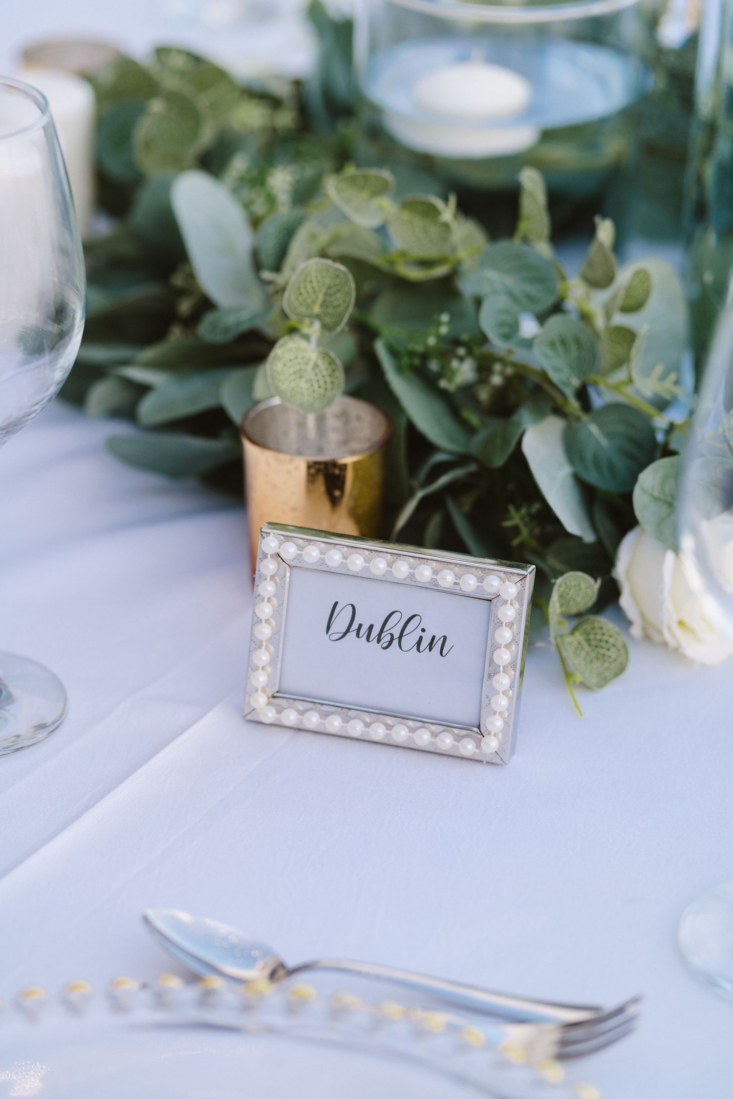 Elegant pearl-framed table card reading 'Dublin' placed on a white tablecloth with eucalyptus greenery decor.