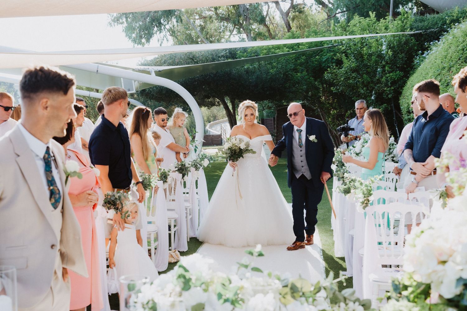 A wedding ceremony outdoors with elegant white floral decorations and guests seated in white chairs.
