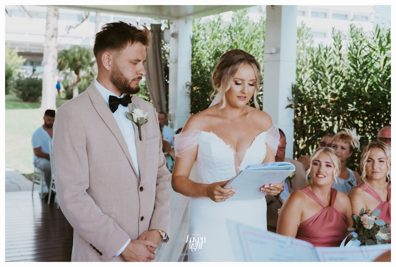 An intimate outdoor wedding scene with guests in coral dresses watching the ceremony.