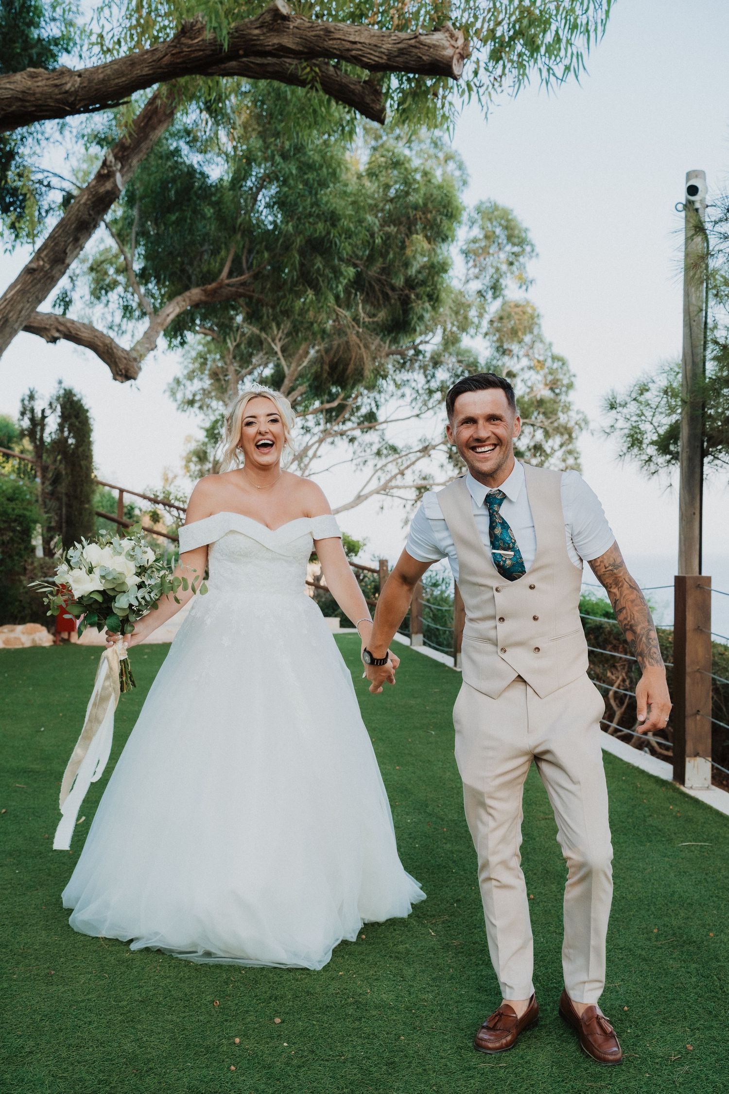 Cheerful bride and groom laugh together on manicured lawn during wedding celebration.
