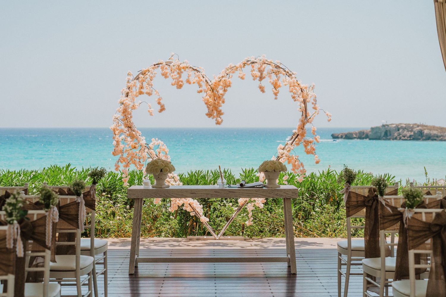 Heart-shaped floral arch overlooking turquoise ocean waters at a beach wedding venue.