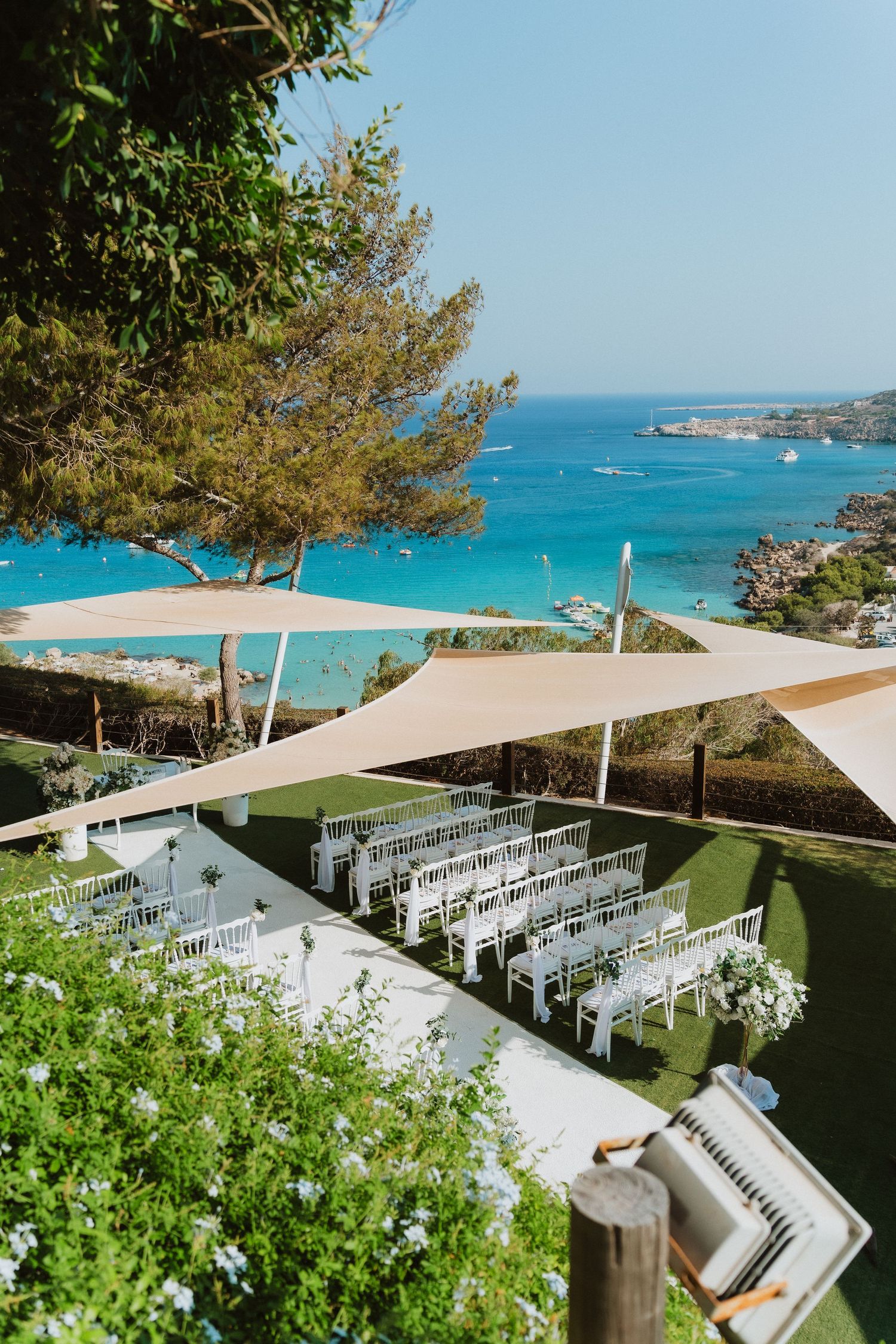 Oceanfront wedding ceremony setup with white chairs and sail canopies overlooking turquoise Mediterranean waters.