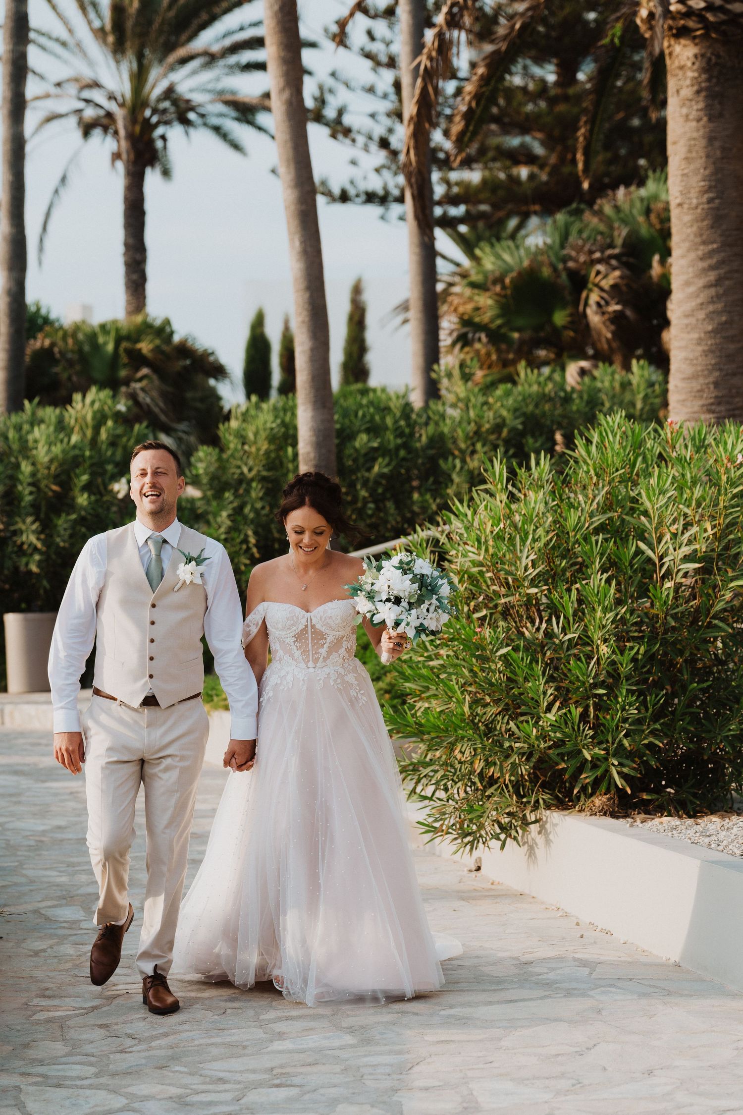 A bride and groom walk together on a tropical garden path with palm trees and Mediterranean plants in the background.