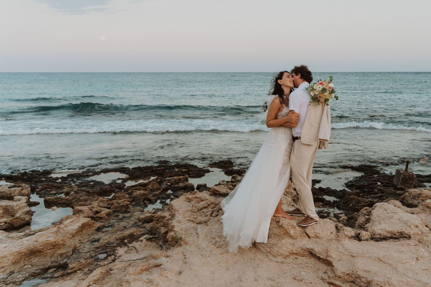 Newlyweds share an intimate moment on rocky coastal terrain during their beach wedding.