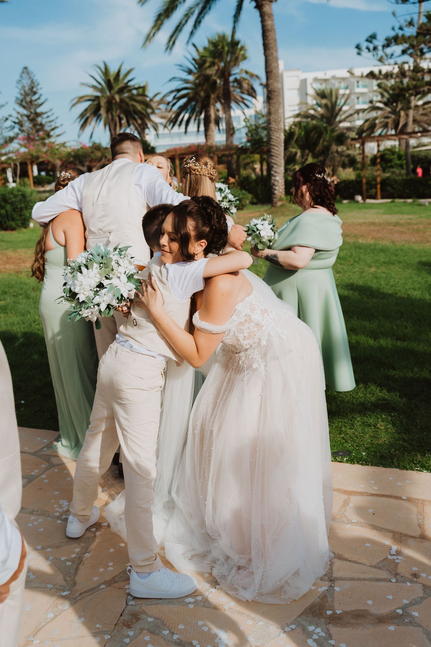 Wedding party in sage green dresses and white attire embrace outdoors with palm trees and greenery in background.