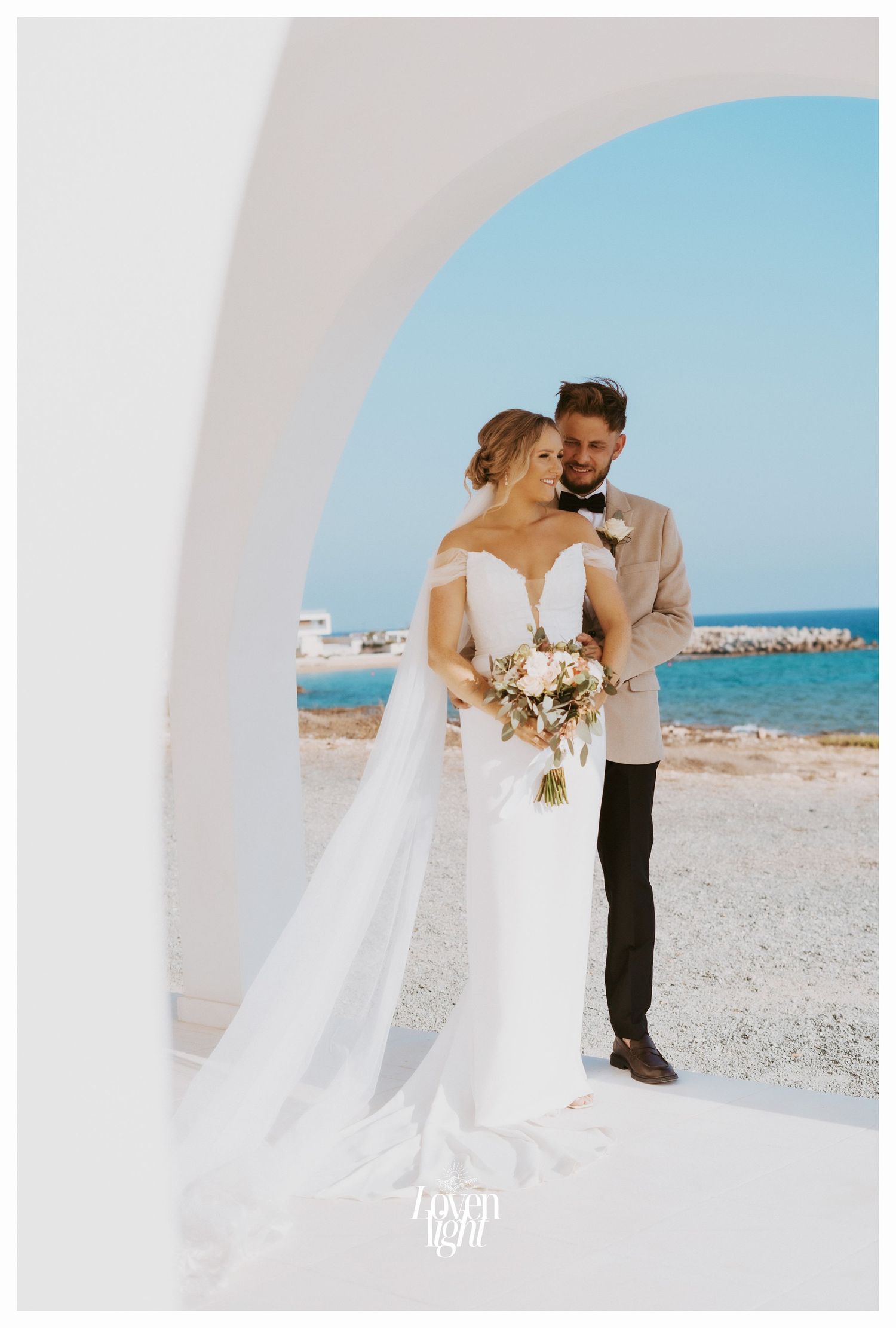 Seaside wedding photograph through white archway with ocean and shoreline visible in background.
