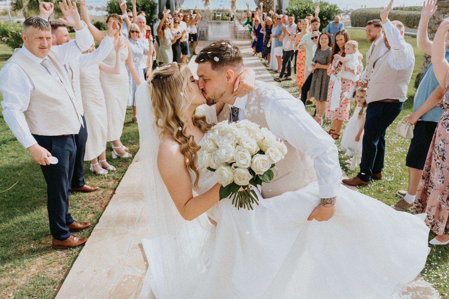 A couple in white wedding attire share a kiss surrounded by cheering guests during an outdoor garden ceremony.
