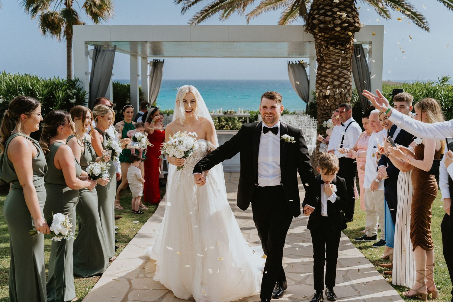 Beachfront wedding exit with guests in formal attire lining the palm-decorated walkway.