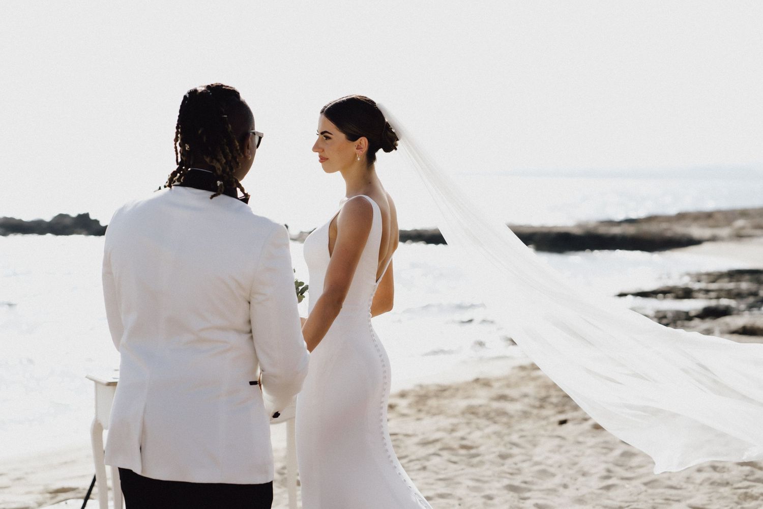 A romantic beach wedding ceremony with flowing white dress and veil against an ocean backdrop.