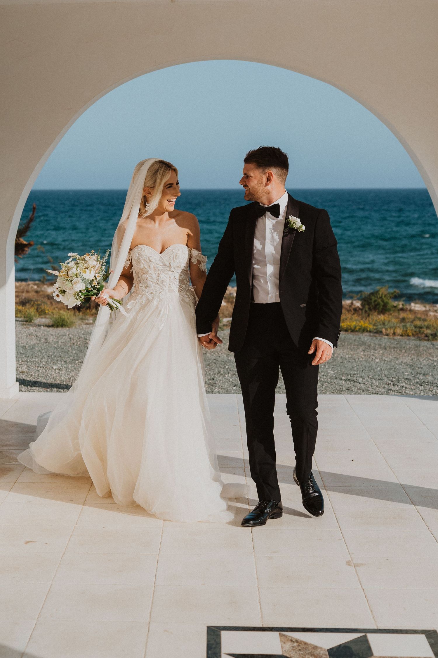 Elegant wedding couple stands under Mediterranean archway overlooking turquoise ocean.