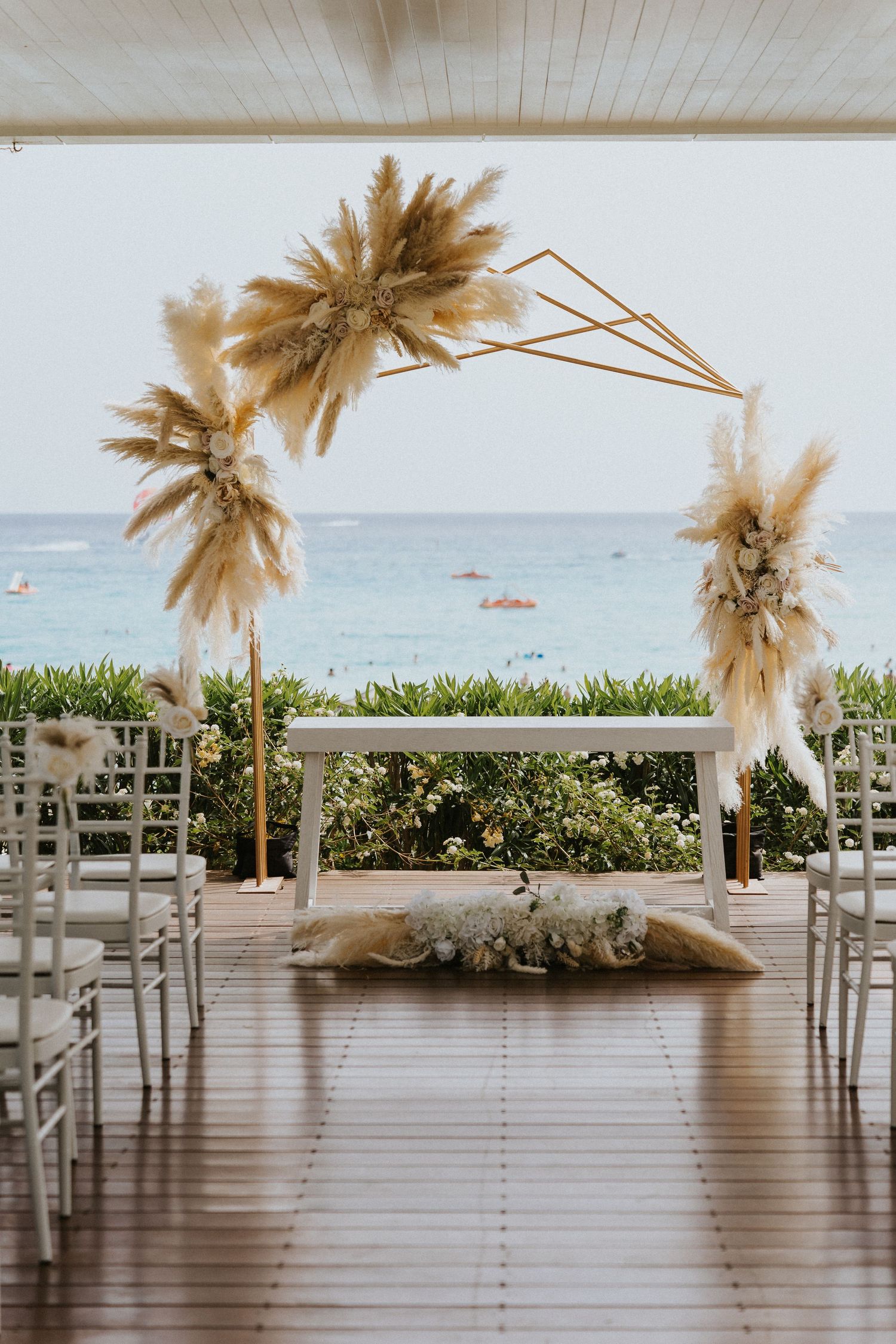 Beachfront wedding ceremony setup with pampas grass arch and white chairs.