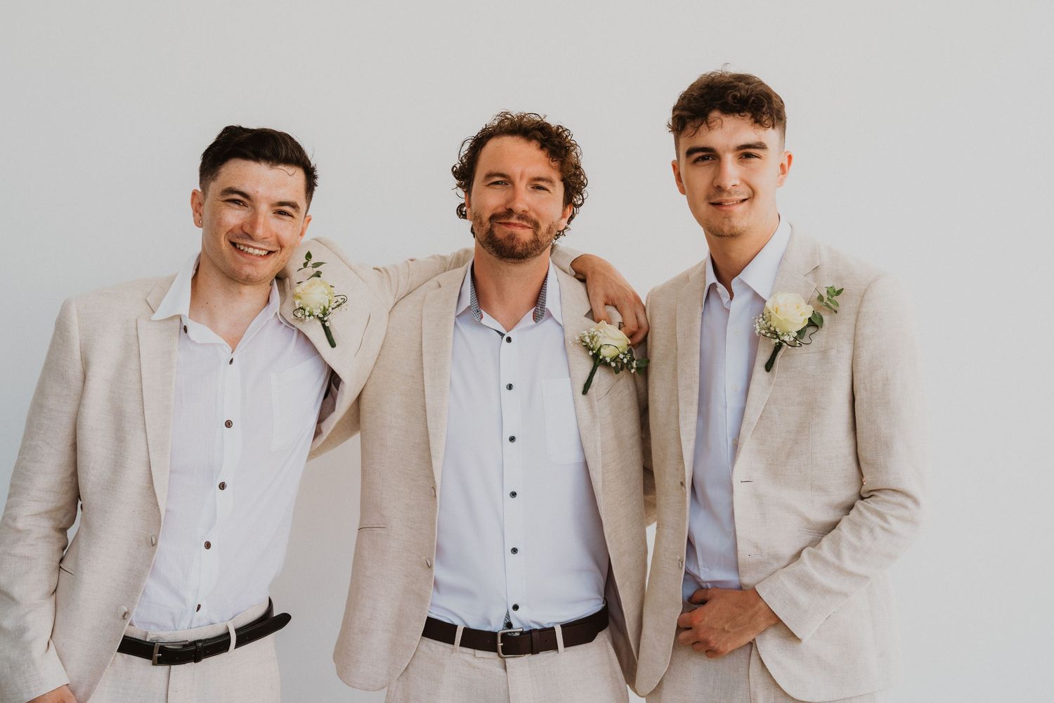 Three men smile in coordinating beige suits with floral boutonnieres for wedding party photo.
