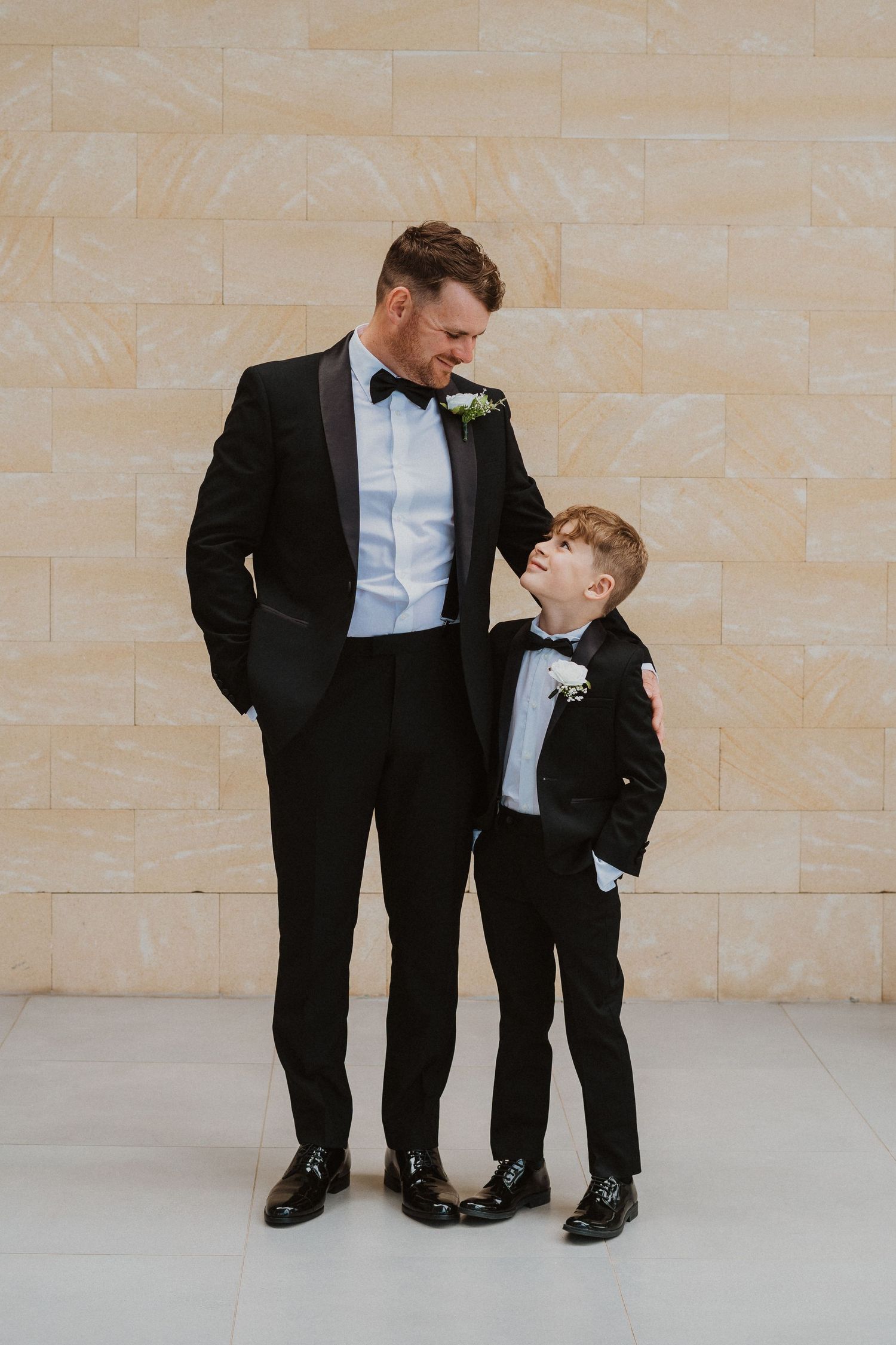 Formal portrait of two people in matching black tuxedos against beige wall.