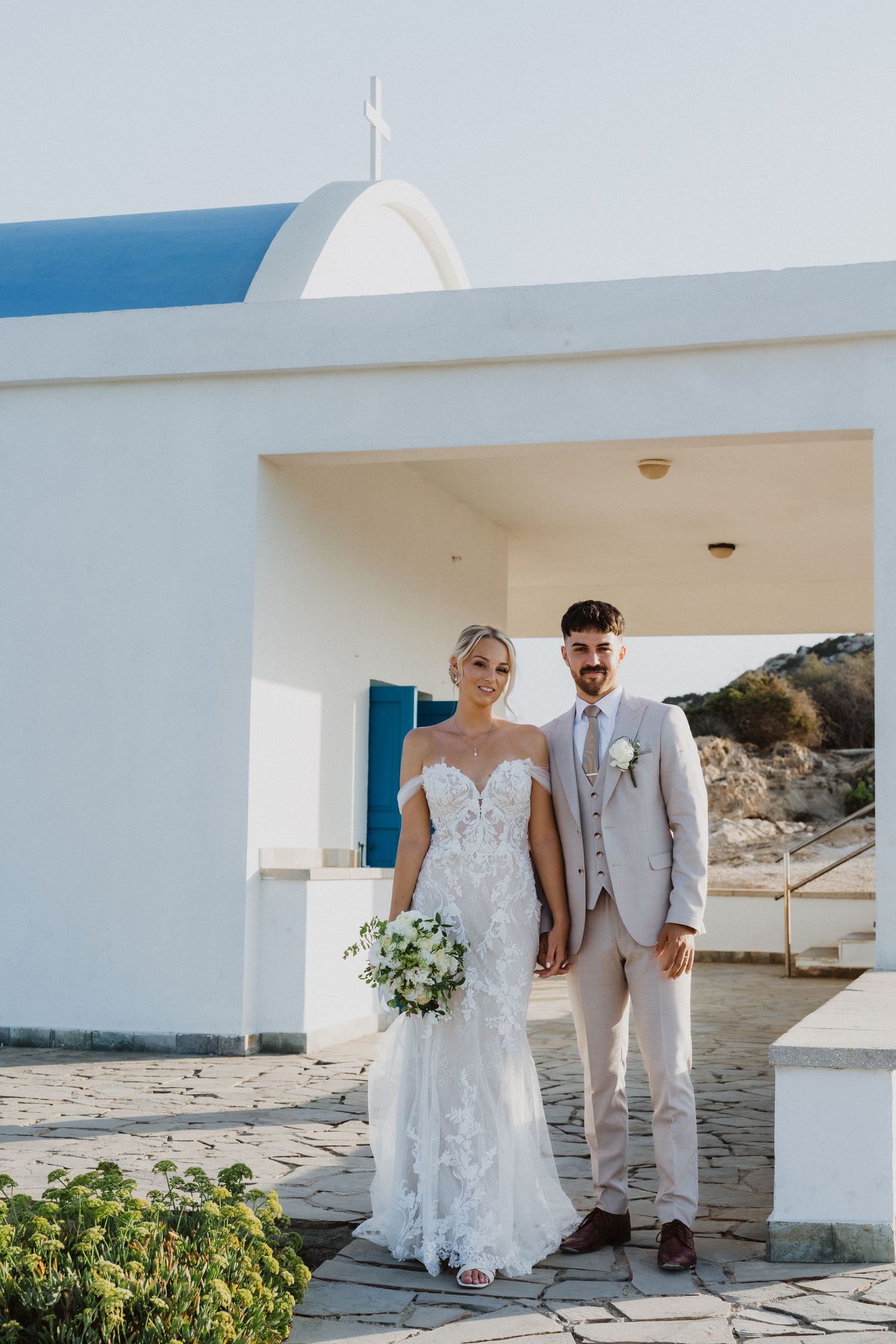 Wedding couple poses against white Mediterranean architecture with blue accents and coastal views.