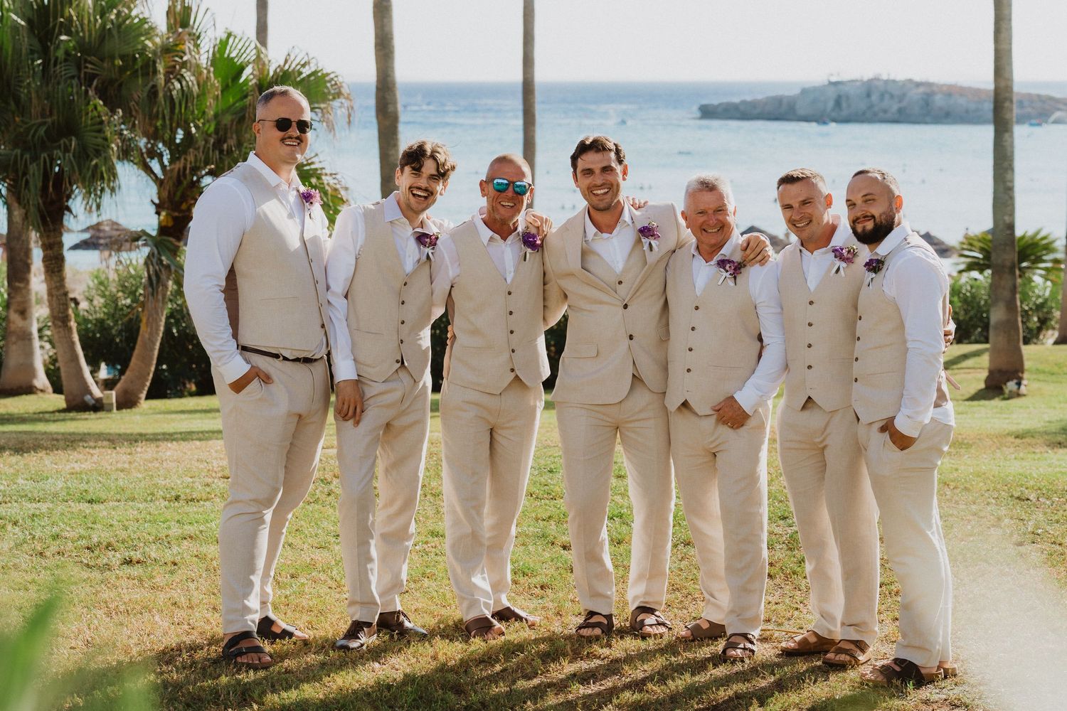 Wedding party members in light colored suits stand in line against tropical beach backdrop.