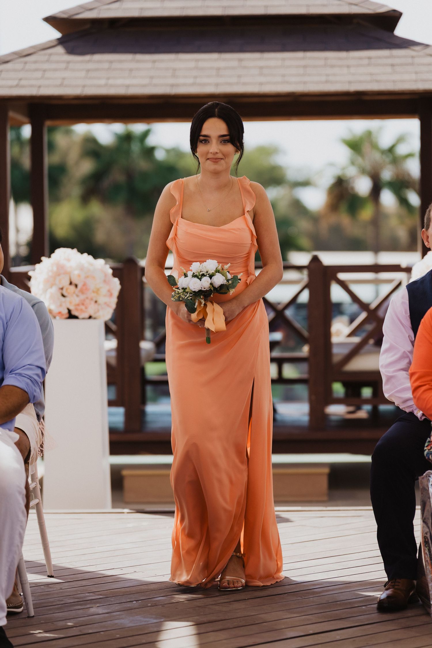 Bridesmaids in coral dresses walk down a wooden deck aisle carrying floral bouquets.