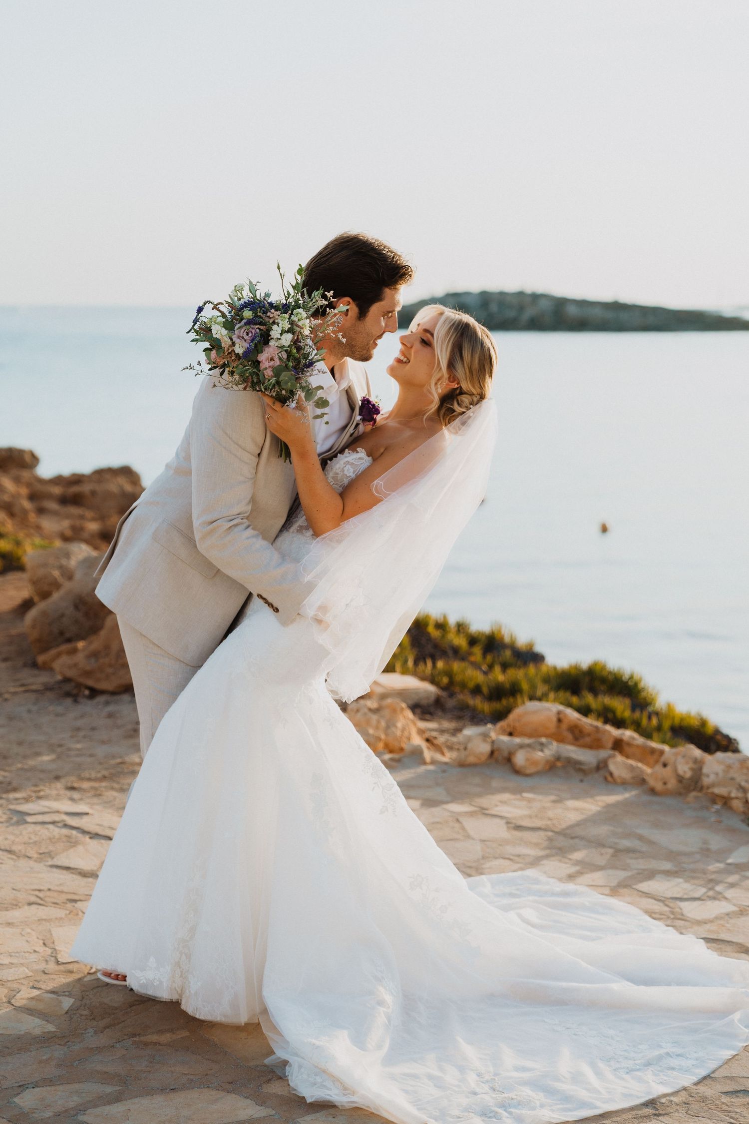 Romantic wedding portrait with couple embracing on coastal cliff at sunset.