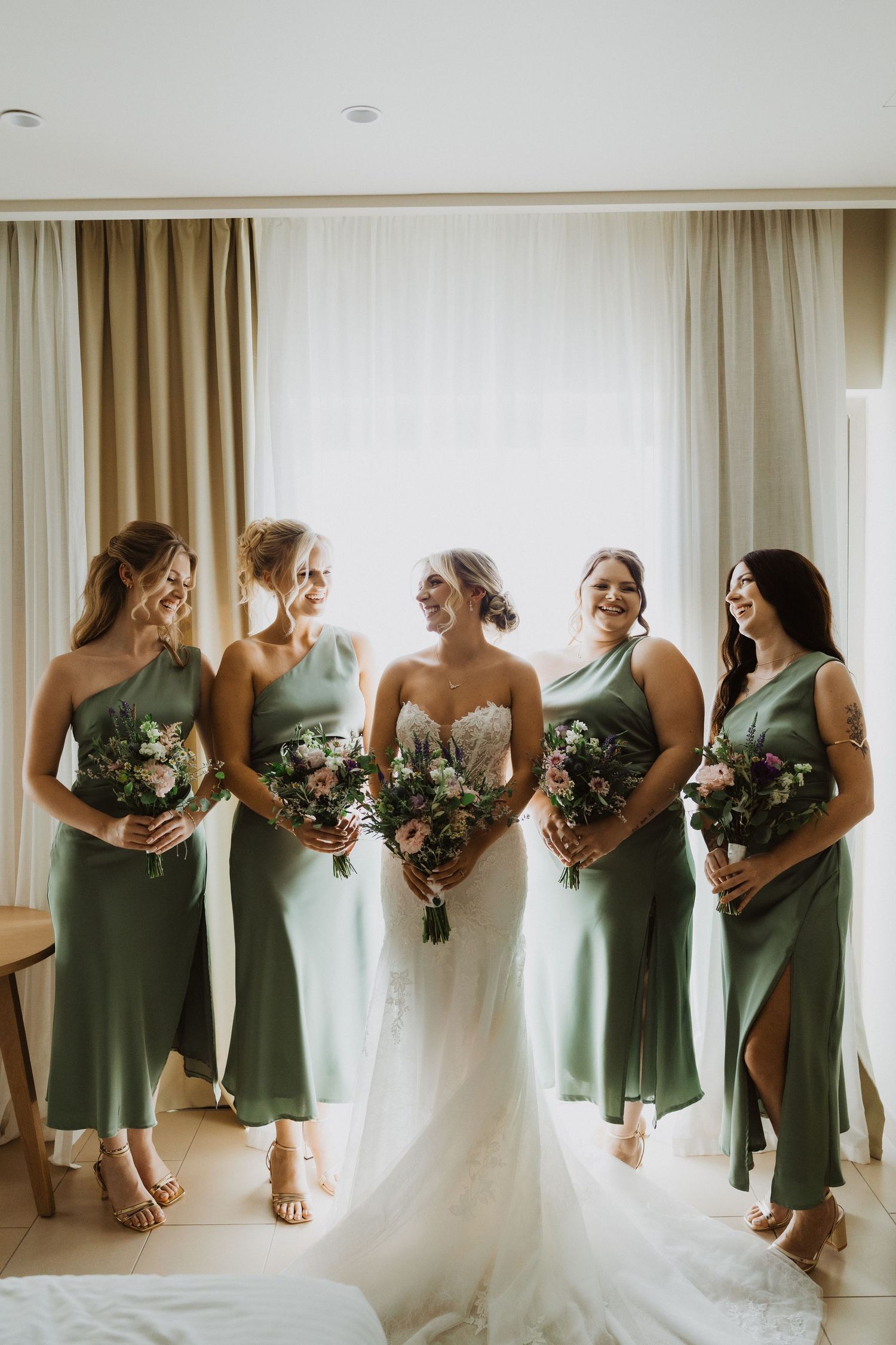 Bridesmaids in matching sage green dresses pose with bride in elegant white gown.