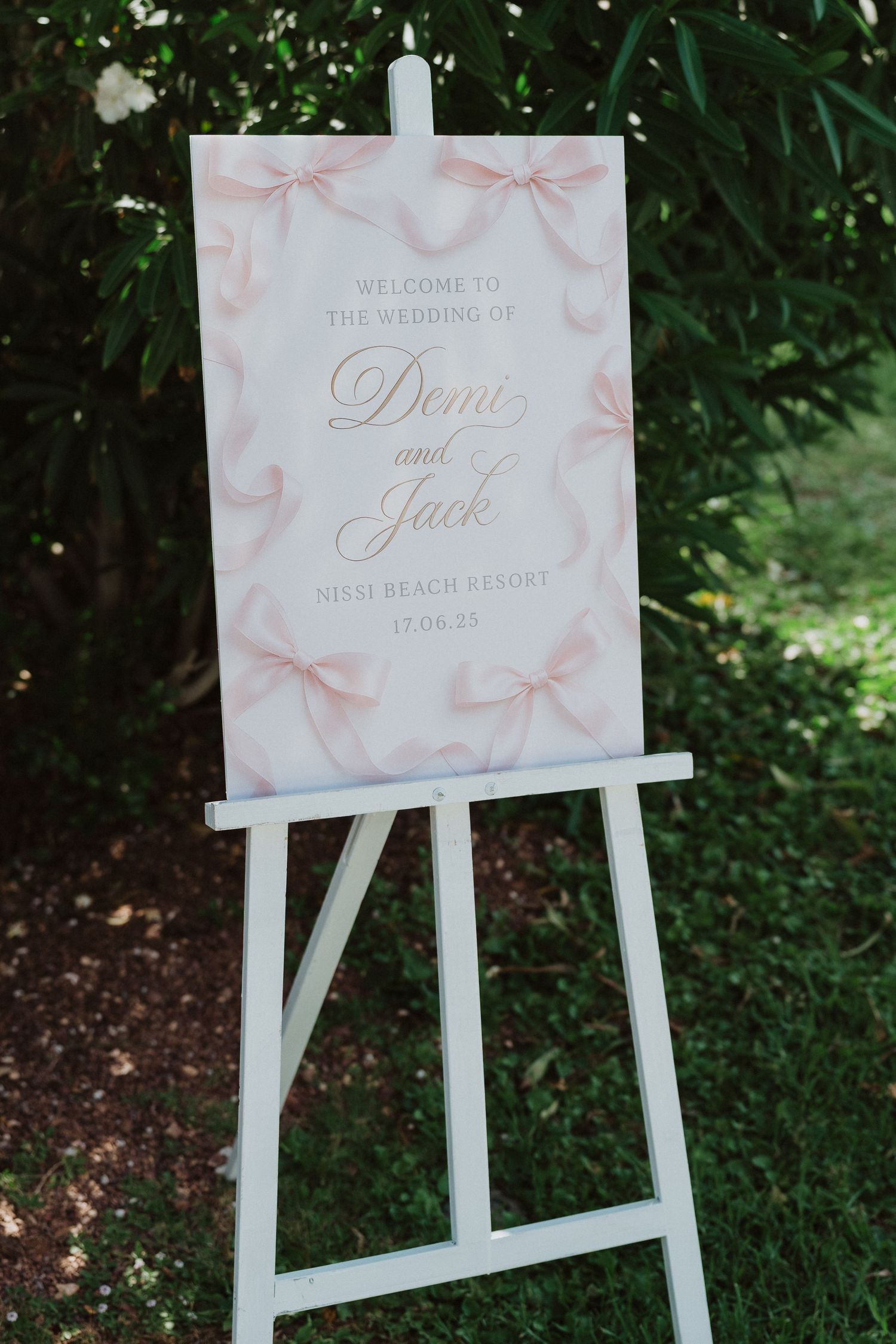White wooden easels display elegant pink and white wedding welcome signs with bow designs in an outdoor garden setting.