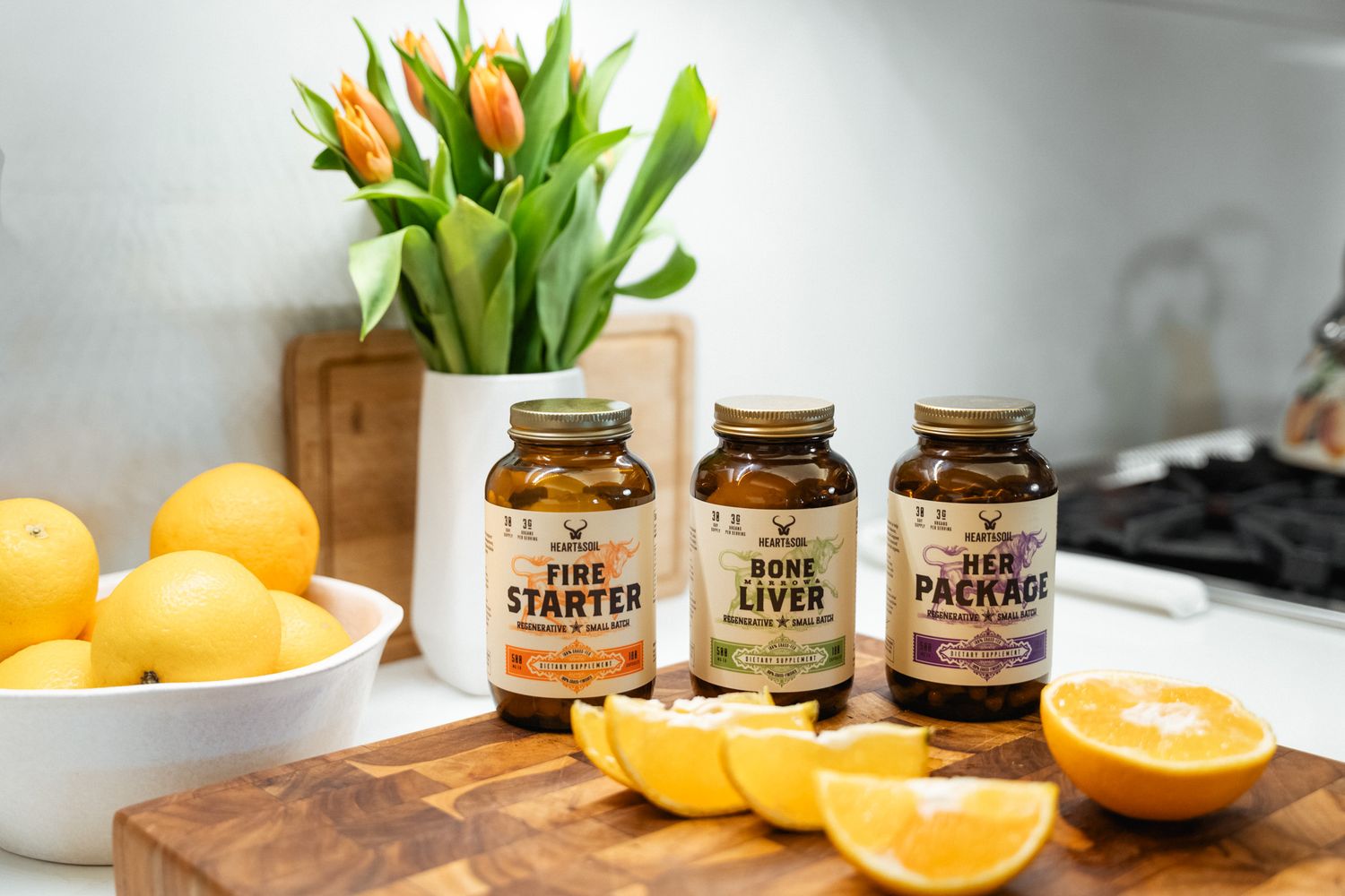 Three supplement bottles labeled Fire Starter, Bone Liver, and Herb Package on a wooden board with fresh oranges and tulips.