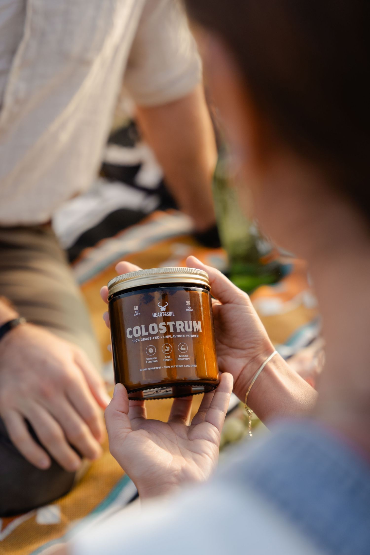 Person holding an amber jar of Colostrum supplement outdoors during a picnic on a colorful blanket.