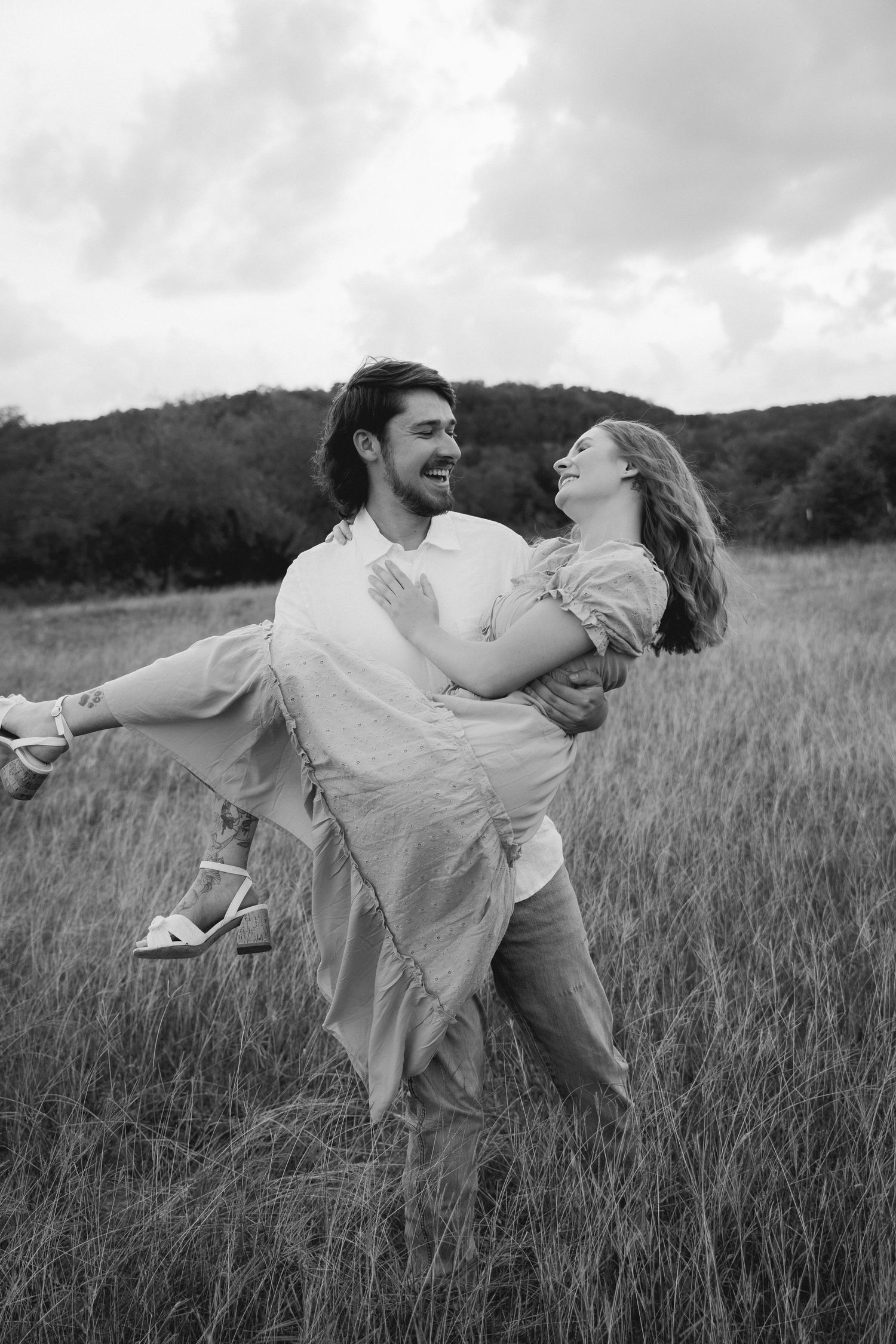 Black and white photo of a joyful couple in a field, man carrying woman in his arms, both laughing outdoors.