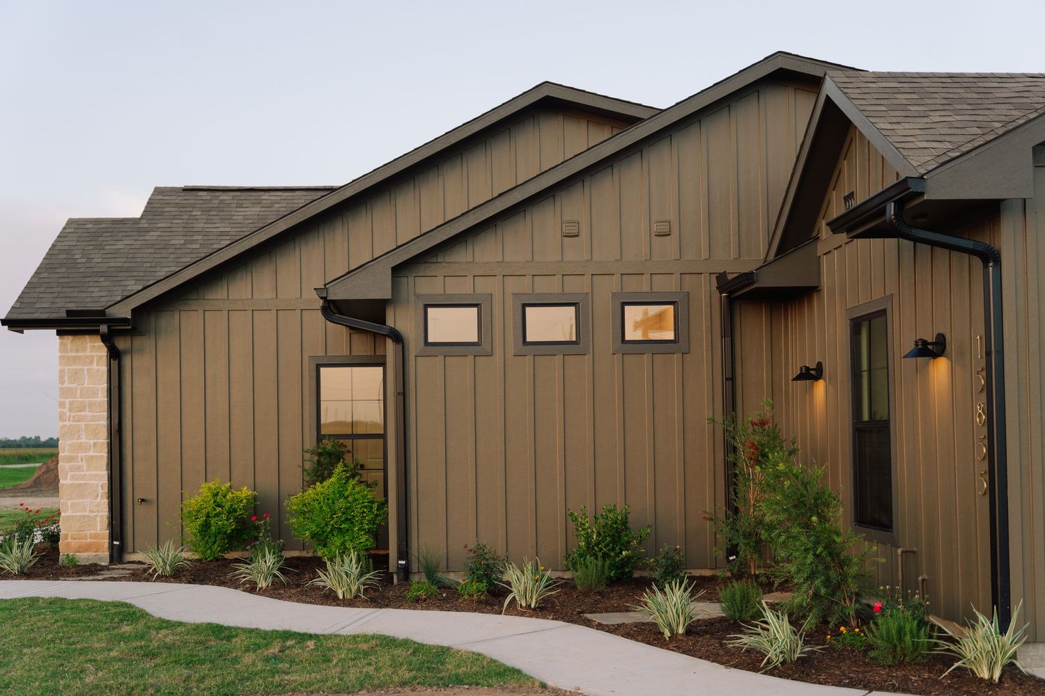 Modern farmhouse exterior with board and batten siding, stone accents, dark trim, and manicured landscaping at dusk.