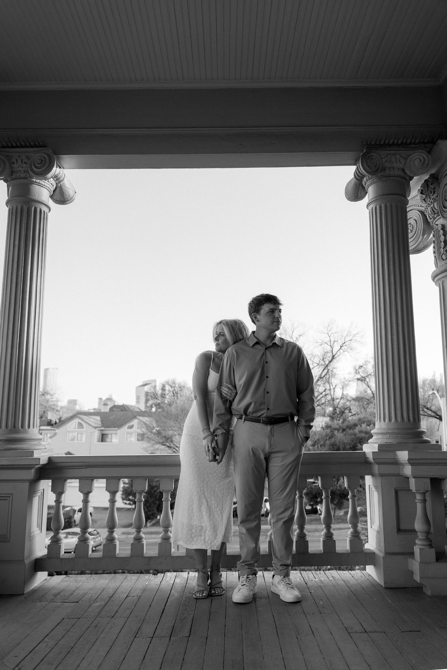 Black and white photo of couple standing between ornate columns on a classic porch with skyline view.