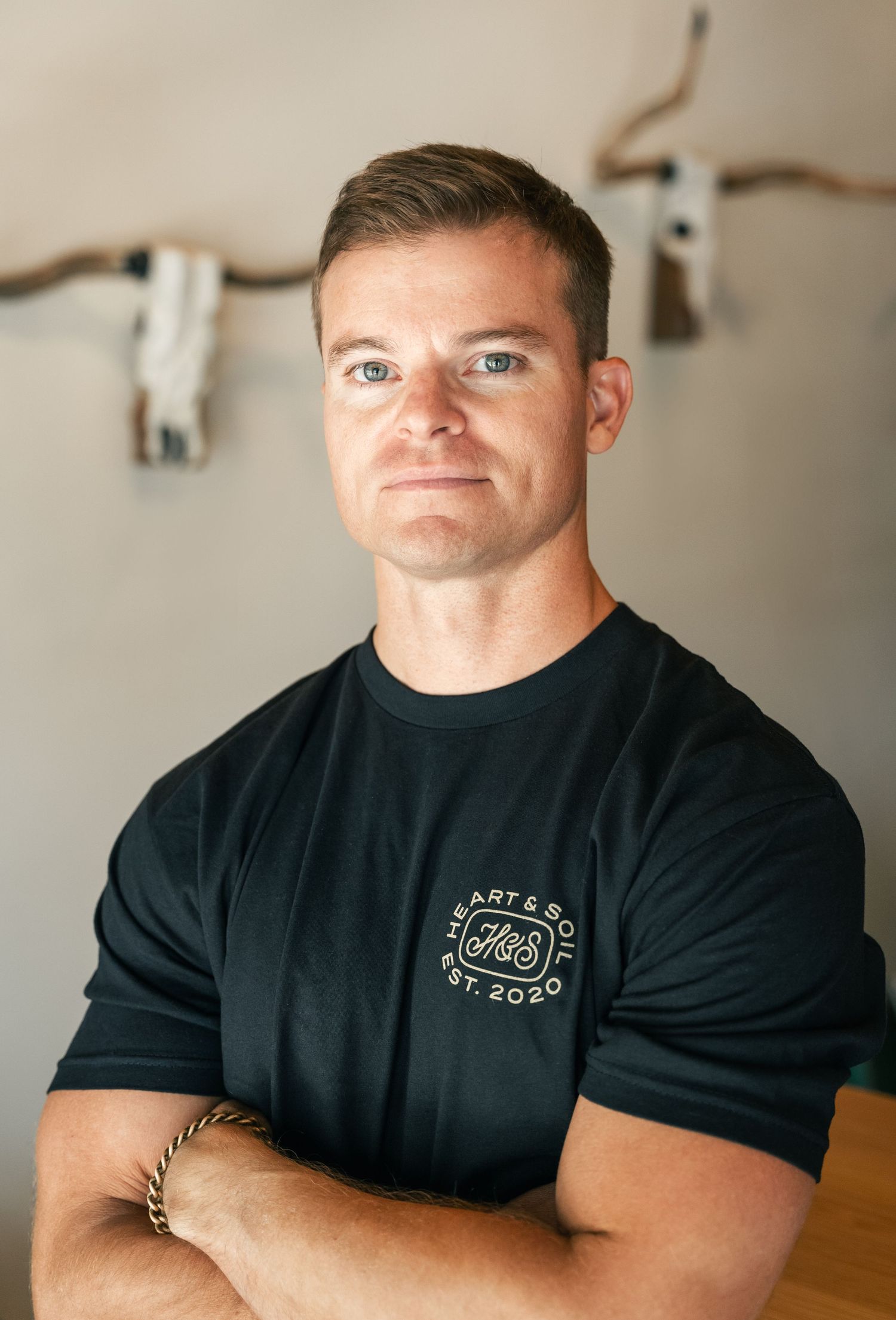 Man in black Heart & Soul Est. 2020 t-shirt poses with arms crossed before wall-mounted longhorn skull decor.