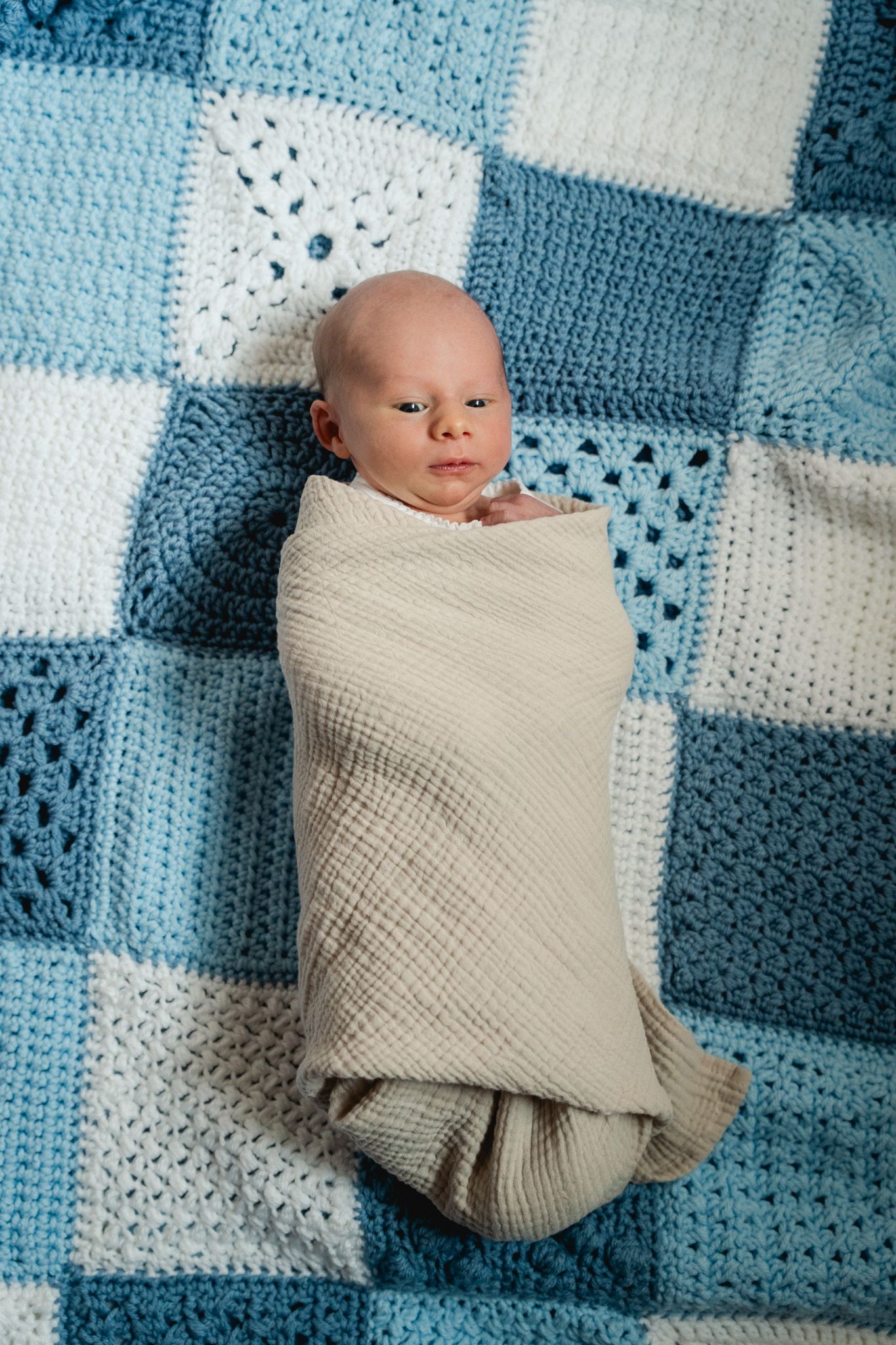 Swaddled newborn baby lying on a blue and white crocheted patchwork blanket, eyes open.