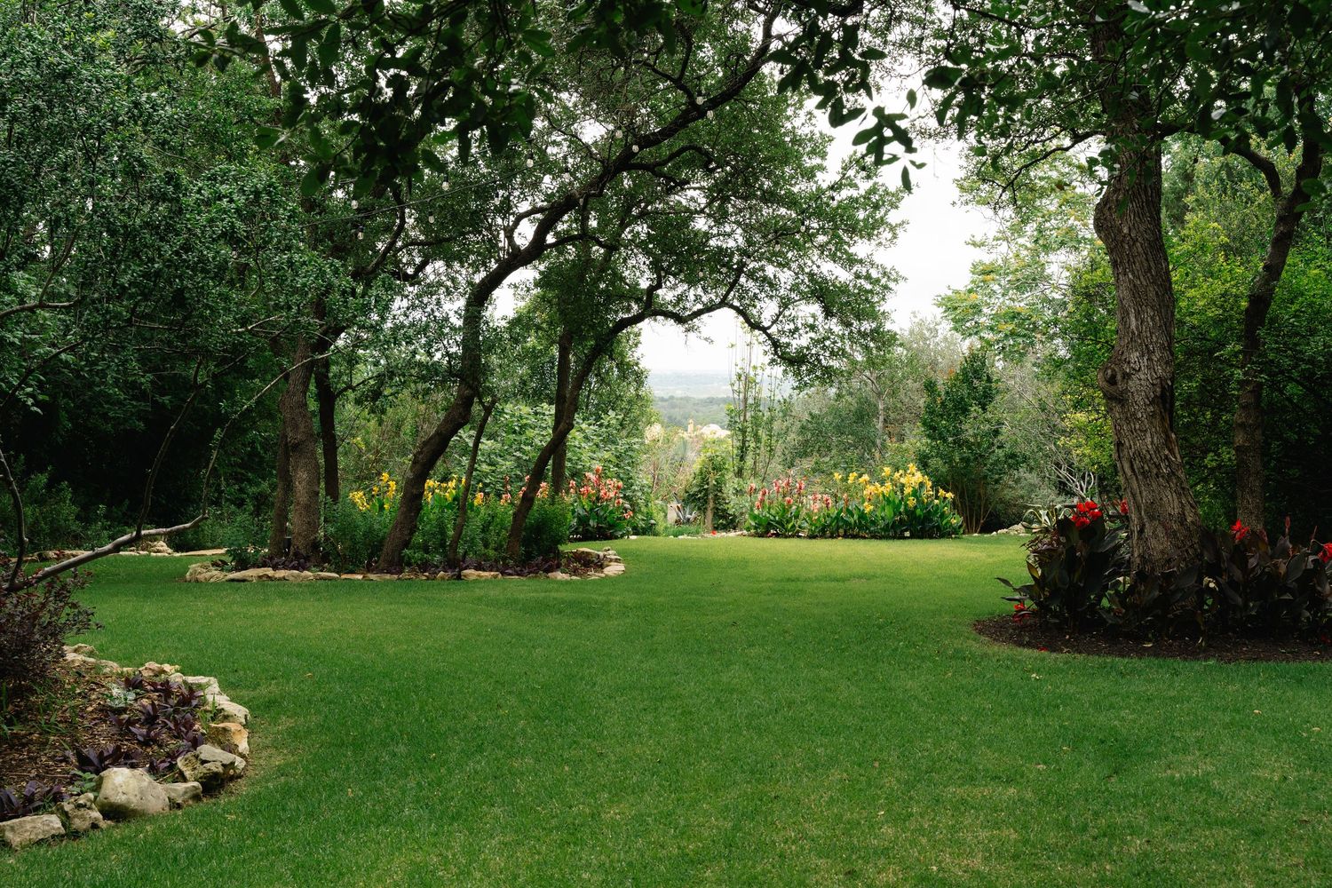 Lush green garden landscape with manicured lawn, mature oak trees, and colorful flower borders in natural setting.