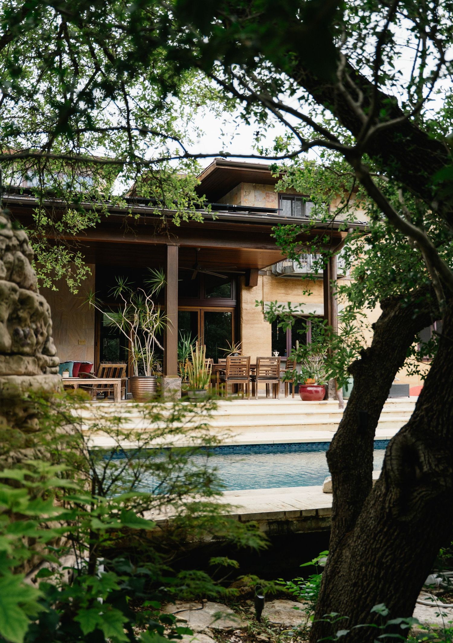 Elegant home with wooden patio furniture and pool viewed through lush green trees in a serene garden setting.