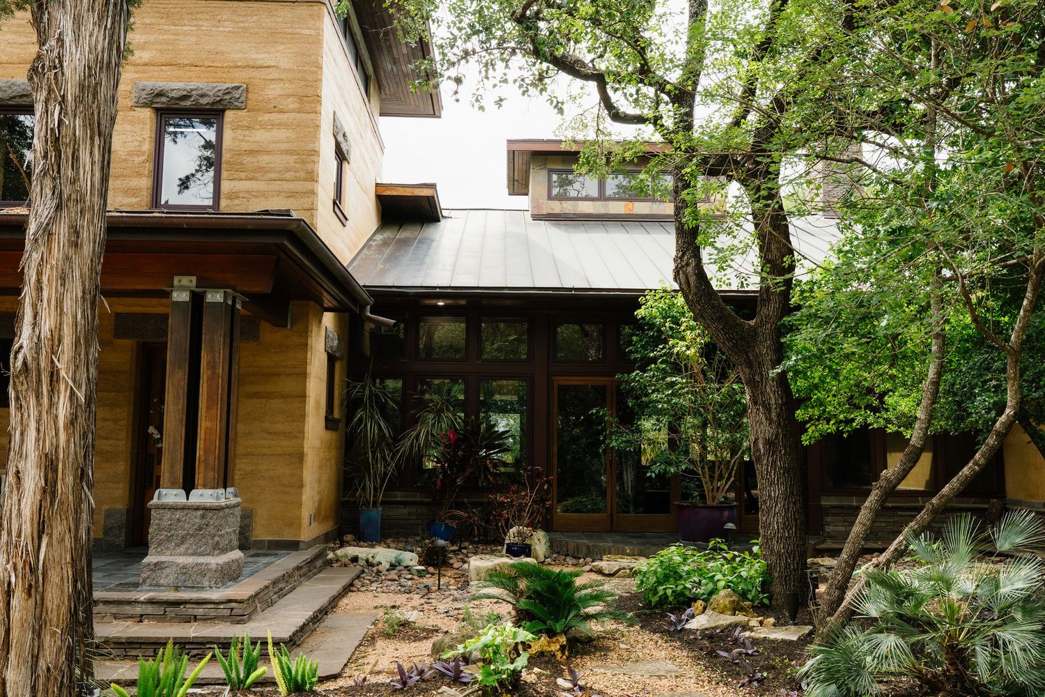 Rammed earth home with metal roof, large glass windows, stone steps, and lush surrounding garden trees.