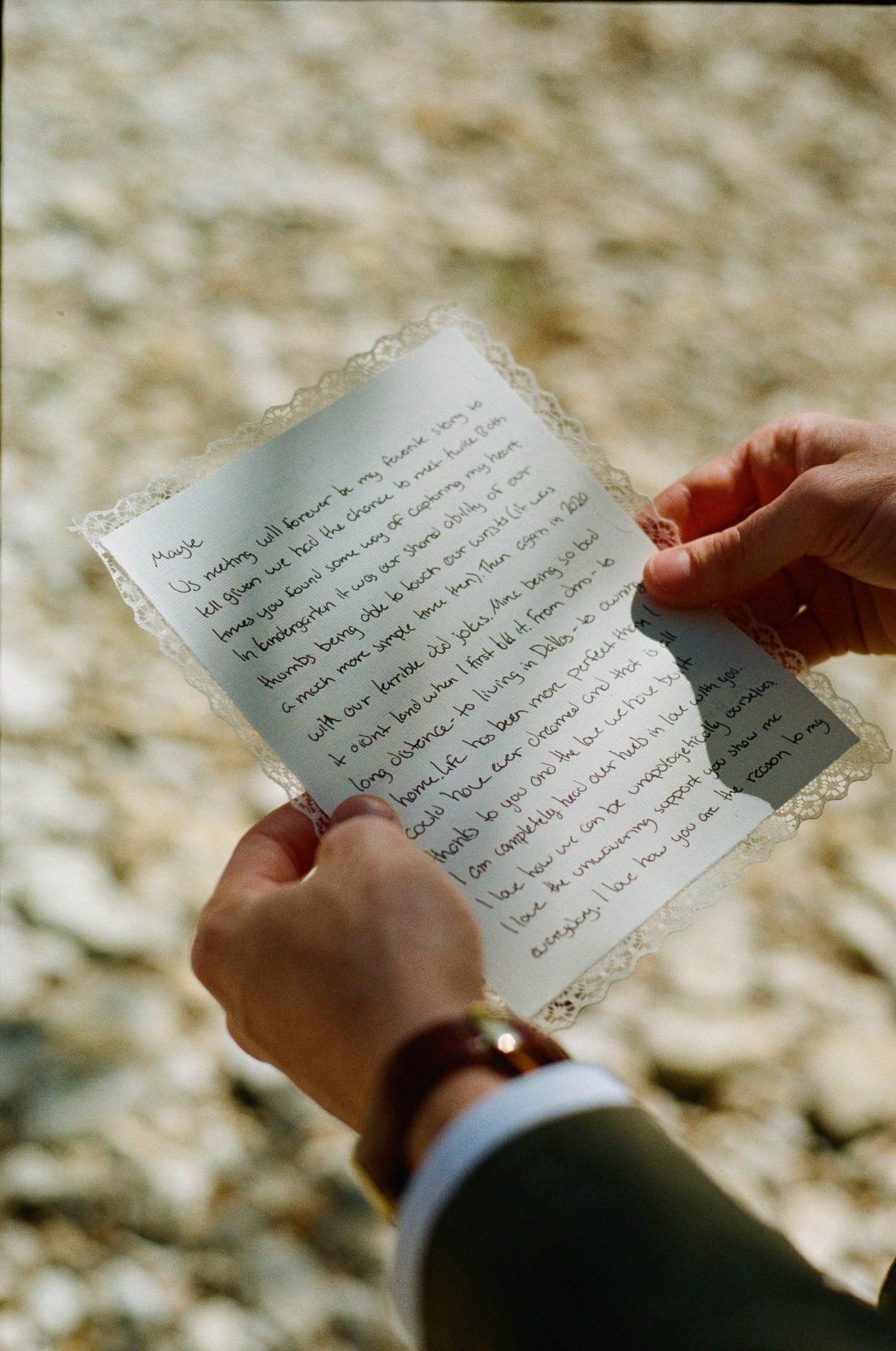 Groom reading handwritten wedding vows letter with lace border outdoors on rocky ground.