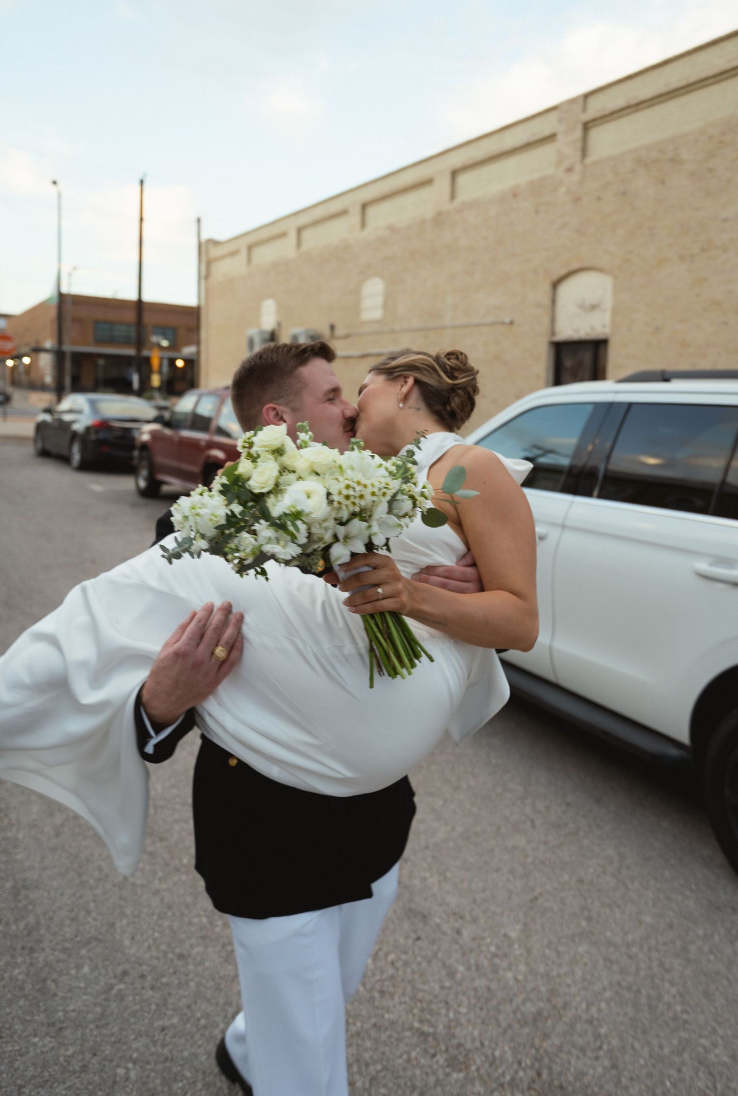 Military groom lifting bride for kiss while holding white floral bouquet.