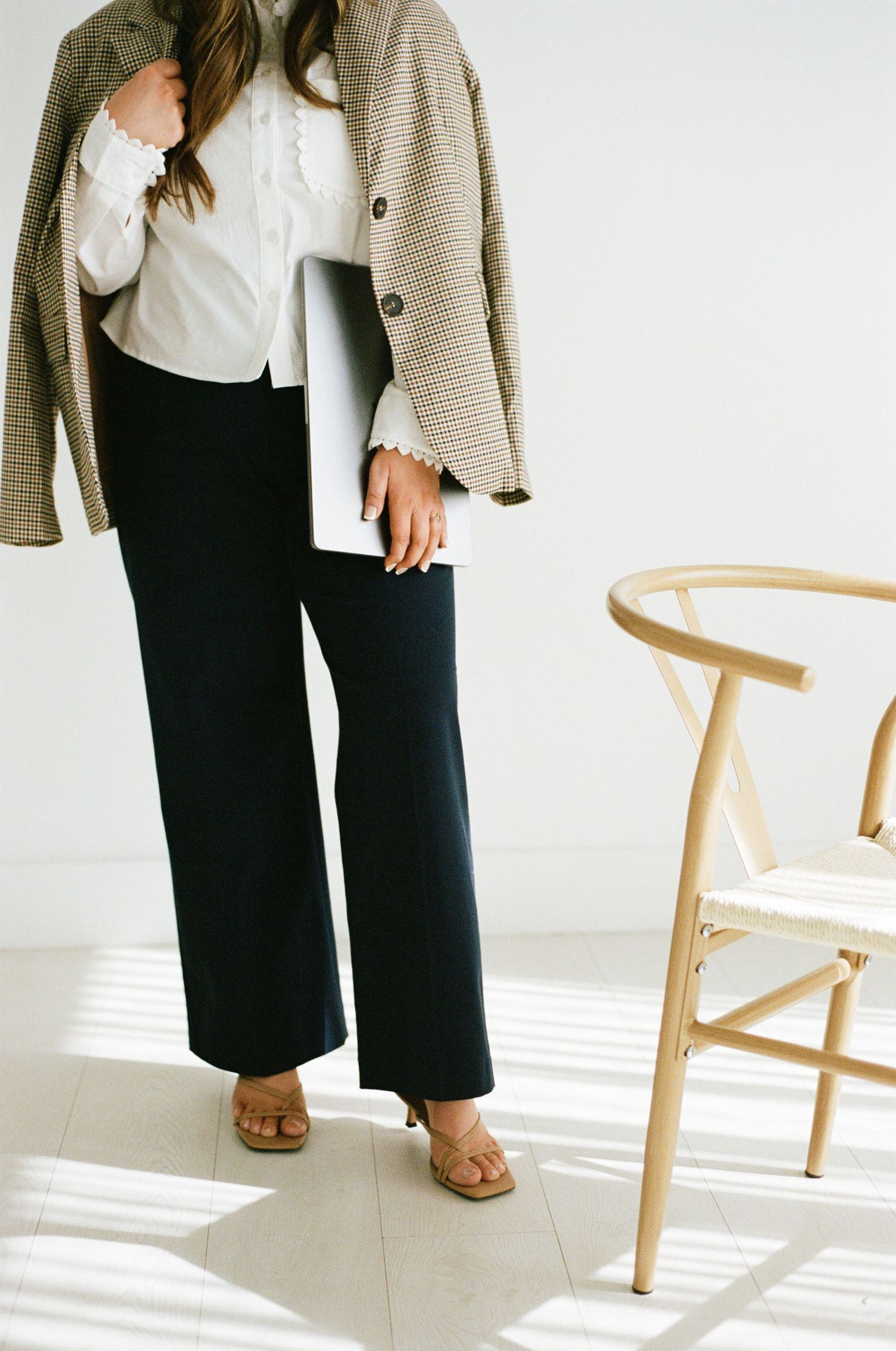 Woman in houndstooth blazer and black wide-leg pants holding a laptop beside a wooden chair.