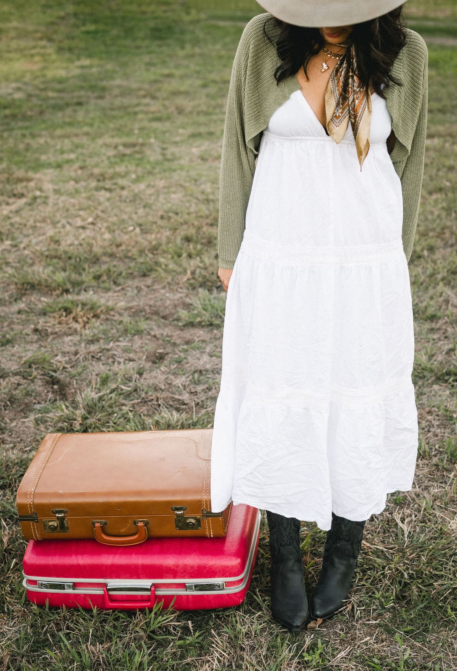 Woman in white maxi dress and green cardigan standing beside stacked vintage suitcases outdoors.