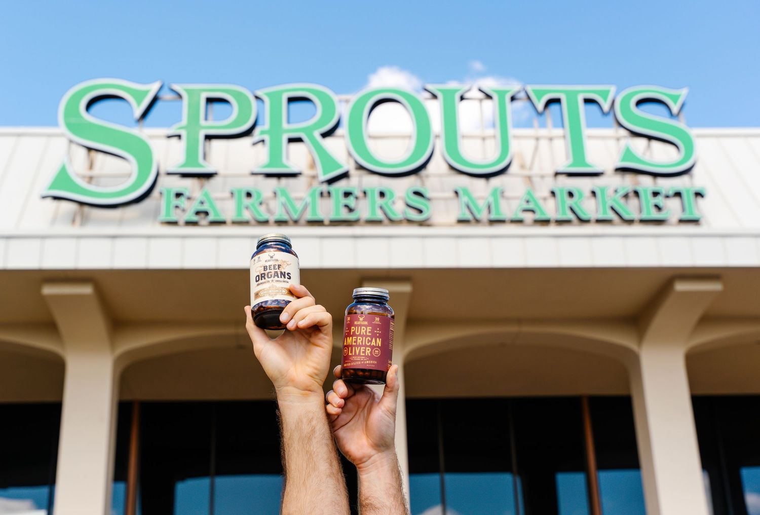Two hands holding supplement jars in front of a Sprouts Farmers Market store exterior.