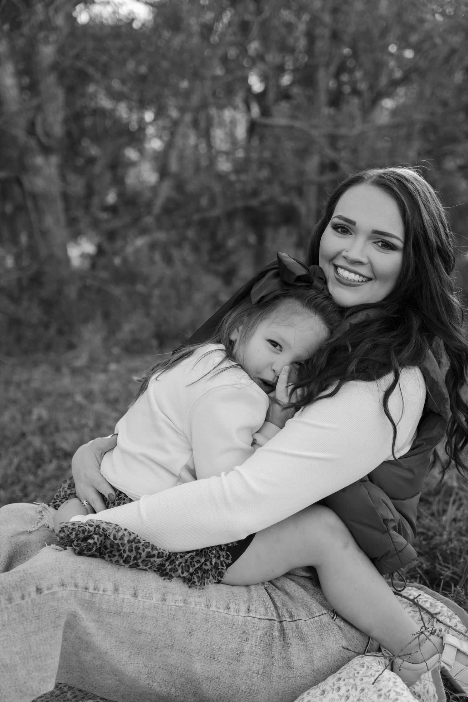 Black and white photo of a smiling mother hugging her young daughter outdoors on a blanket.