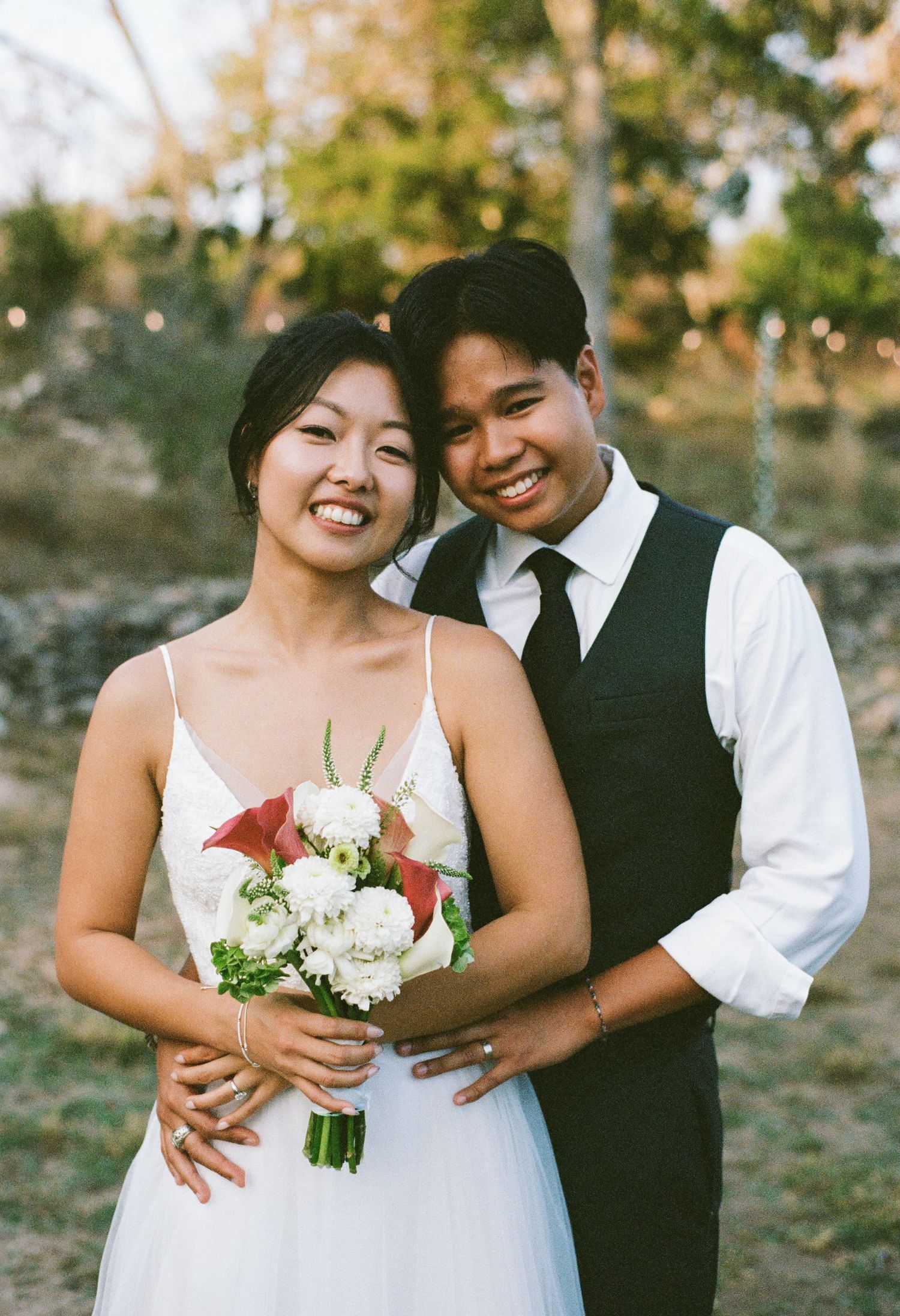 Smiling Asian bride and groom posing with wedding bouquet at outdoor ceremony.