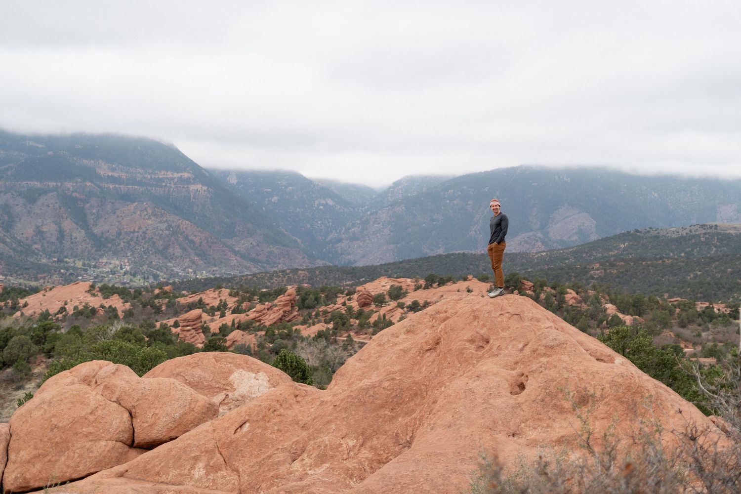 Hiker standing atop red rock formation overlooking vast mountain valley with misty peaks and green landscape below.