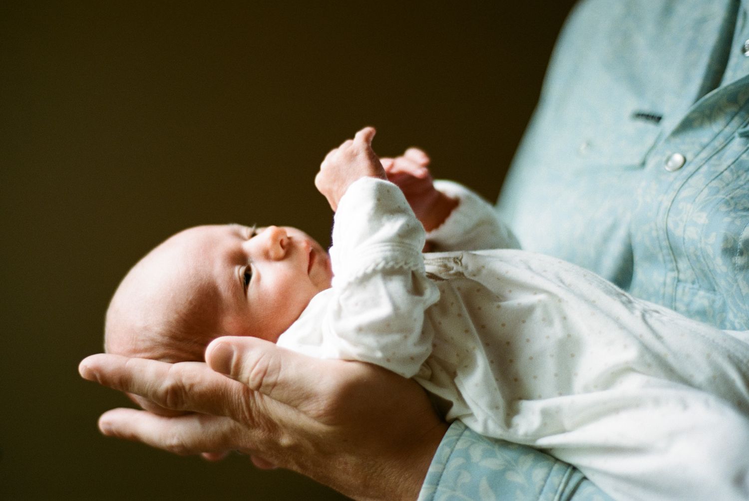 A newborn baby resting peacefully in the gentle hands of an adult, wearing a white onesie against a dark background.