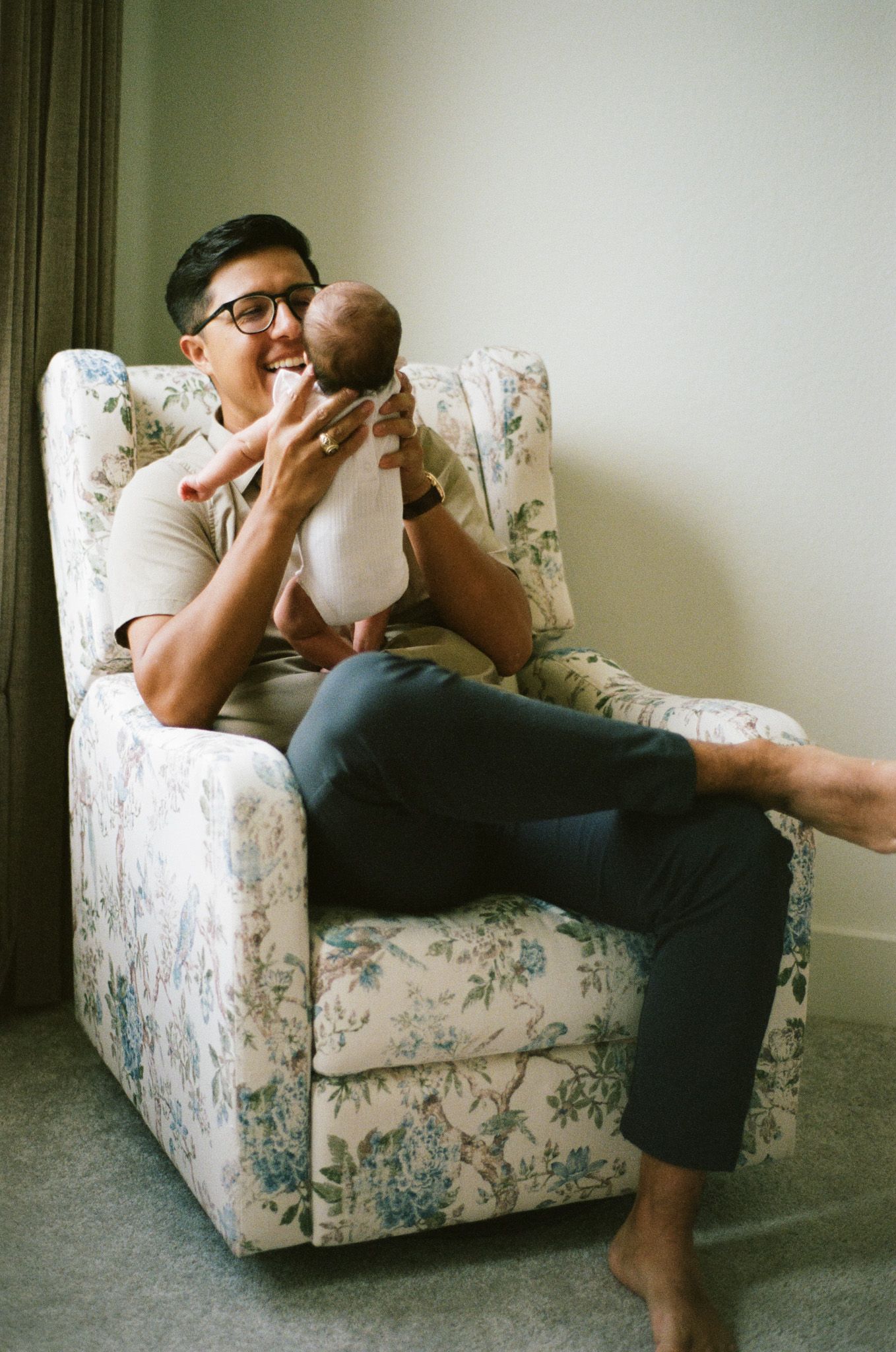 Smiling father in glasses sitting in floral armchair lovingly lifting newborn baby up to his face.