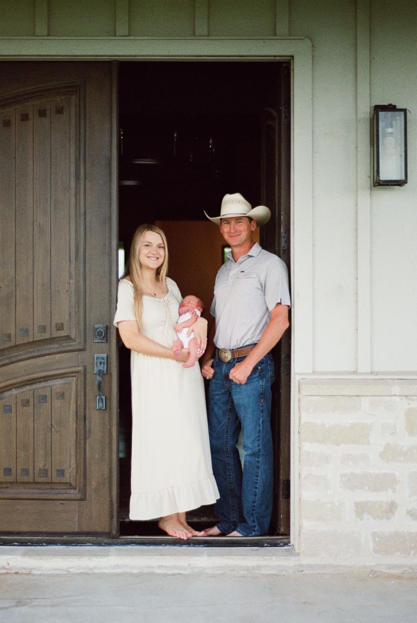 Young couple standing in doorway holding newborn baby, man wearing cowboy hat and jeans, woman in white dress.