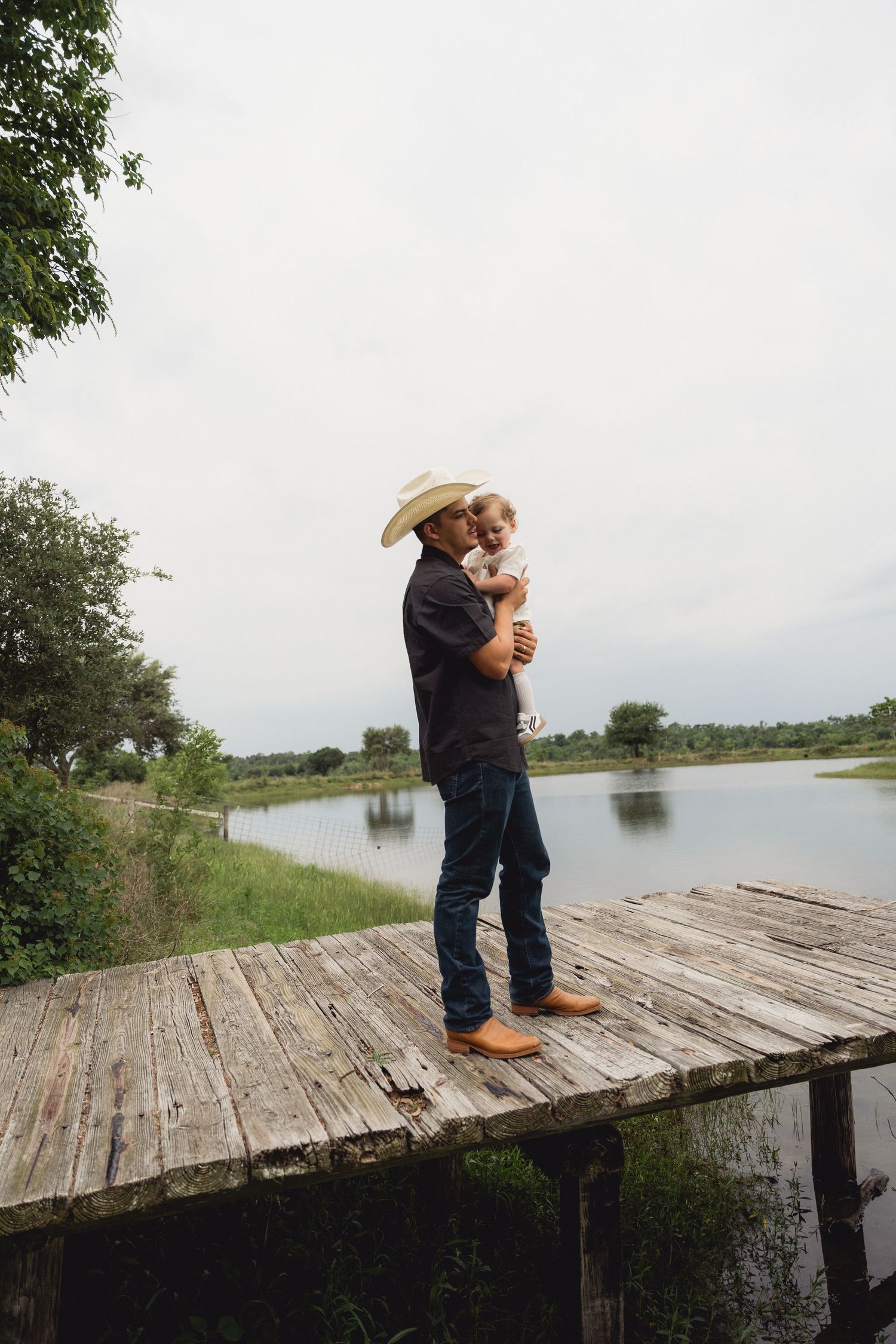 A father in a cowboy hat tenderly holds his baby while standing on a rustic lakeside dock.