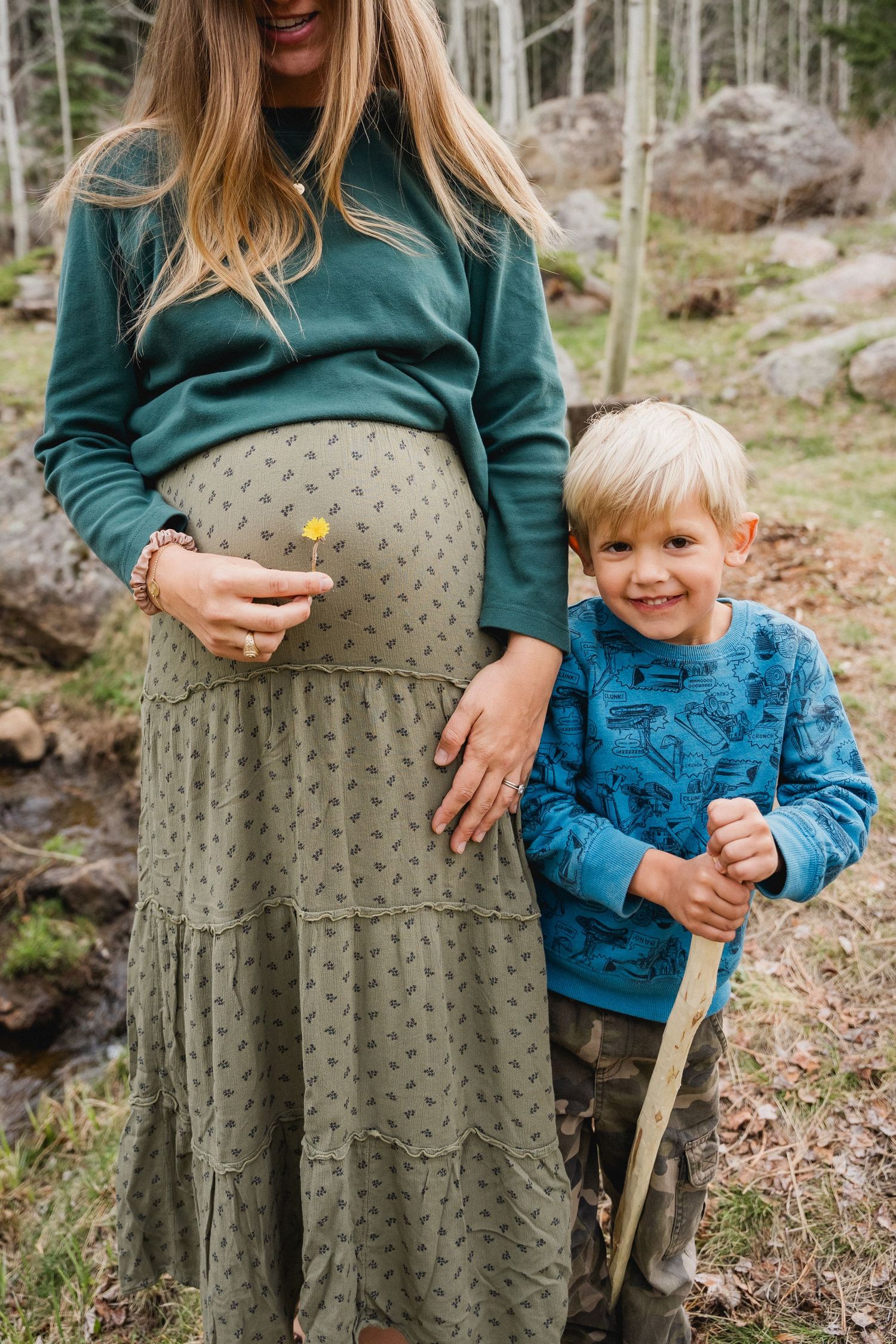 Pregnant mother in green outfit standing with smiling young blonde boy in forest setting.