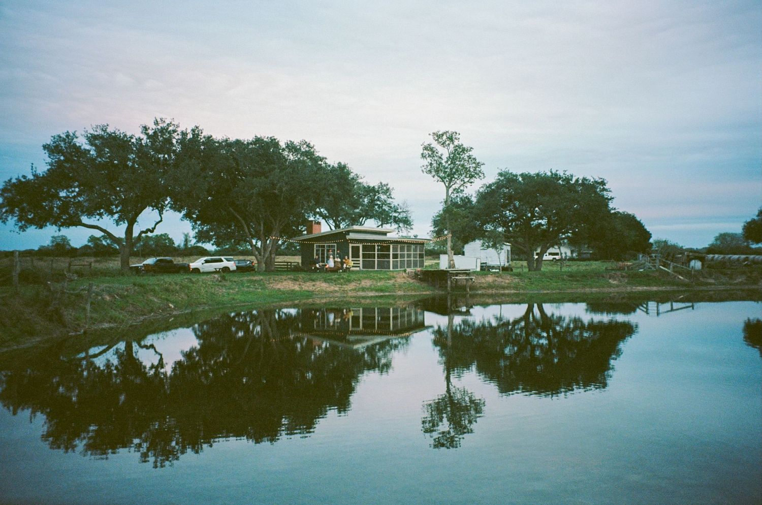 Peaceful lakeside reflection of trees and a building under a soft evening sky.